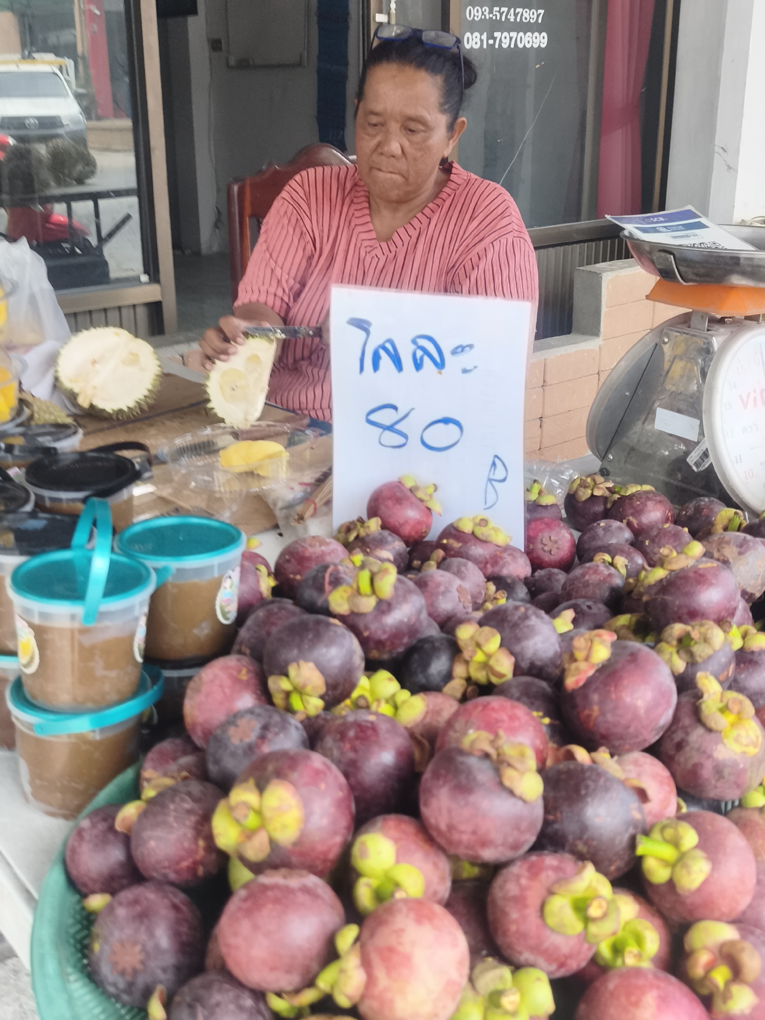 A woman is preparing fruit at a market stall. She is wearing a pink striped shirt and glasses on top of her head. In front of her are several ripe mangosteens in a green basket, and a sliced durian. There are also two plastic containers with brown liquid, possibly juice or a sauce. A scale is visible to the right of the woman, and a sign with Thai writing and the number "80" is placed on the table. The scene appears to be outdoors, under daylight, with a white car visible in the background through a glass window. The woman's expression is focused as she cuts the durian. Visible text includes phone numbers: "093-5747897" and "081-7970699".