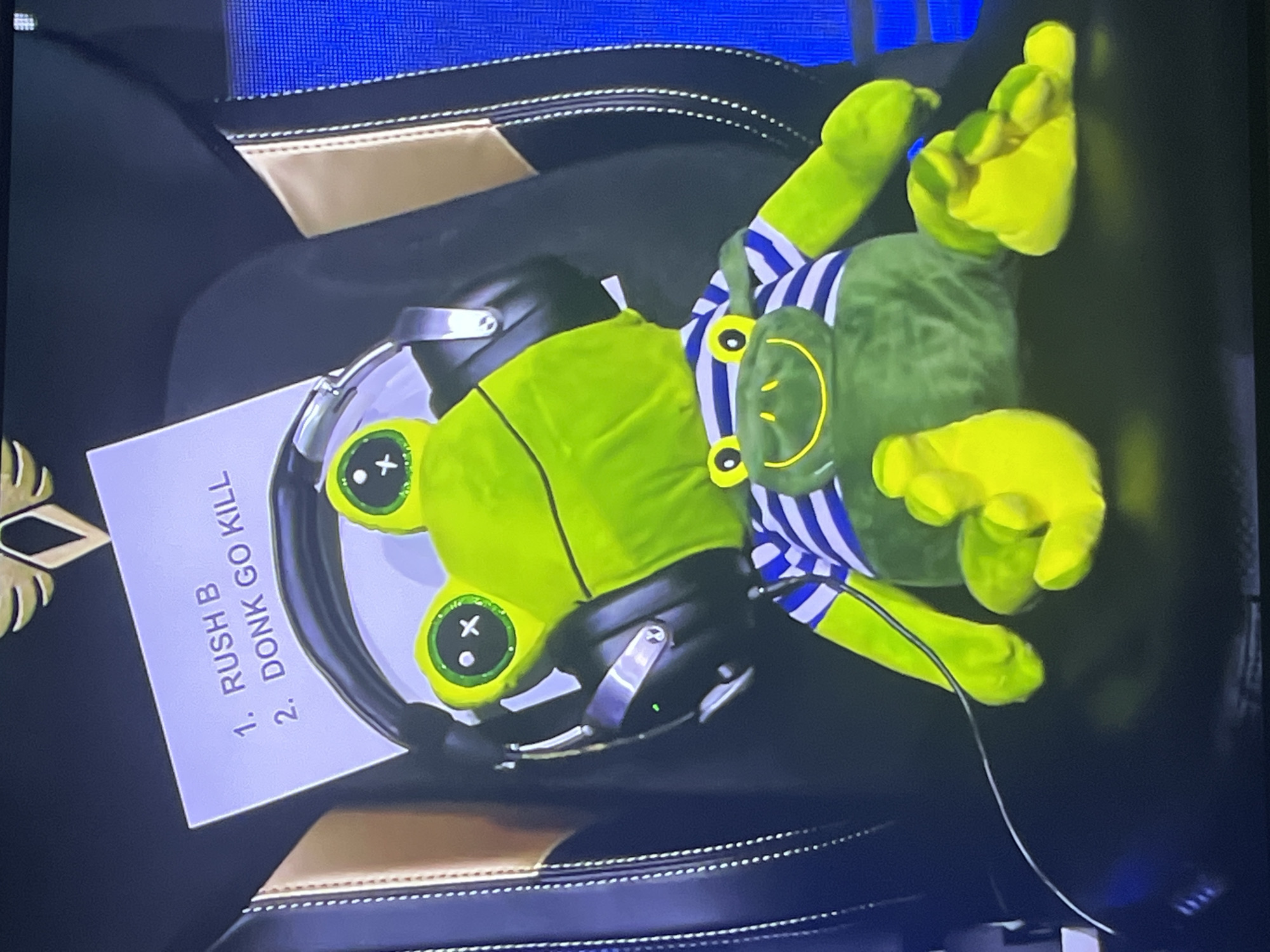 The image shows a plush toy frog wearing a blue and white striped shirt, resting on a black and gold seat. The frog is lying on its back, with its limbs spread out. A white piece of paper is placed under the frog's head, with the handwritten text "1. RUSH B" and "2. DONK GO KILL" visible. Headphones are placed on top of the paper and partially over the frog's face. The background is blurred but appears to be an indoor setting, possibly a room with blue accents. The lighting suggests it could be daytime or artificial light. The overall scene appears to be staged, with the frog and props arranged deliberately. There are no people visible in the image.