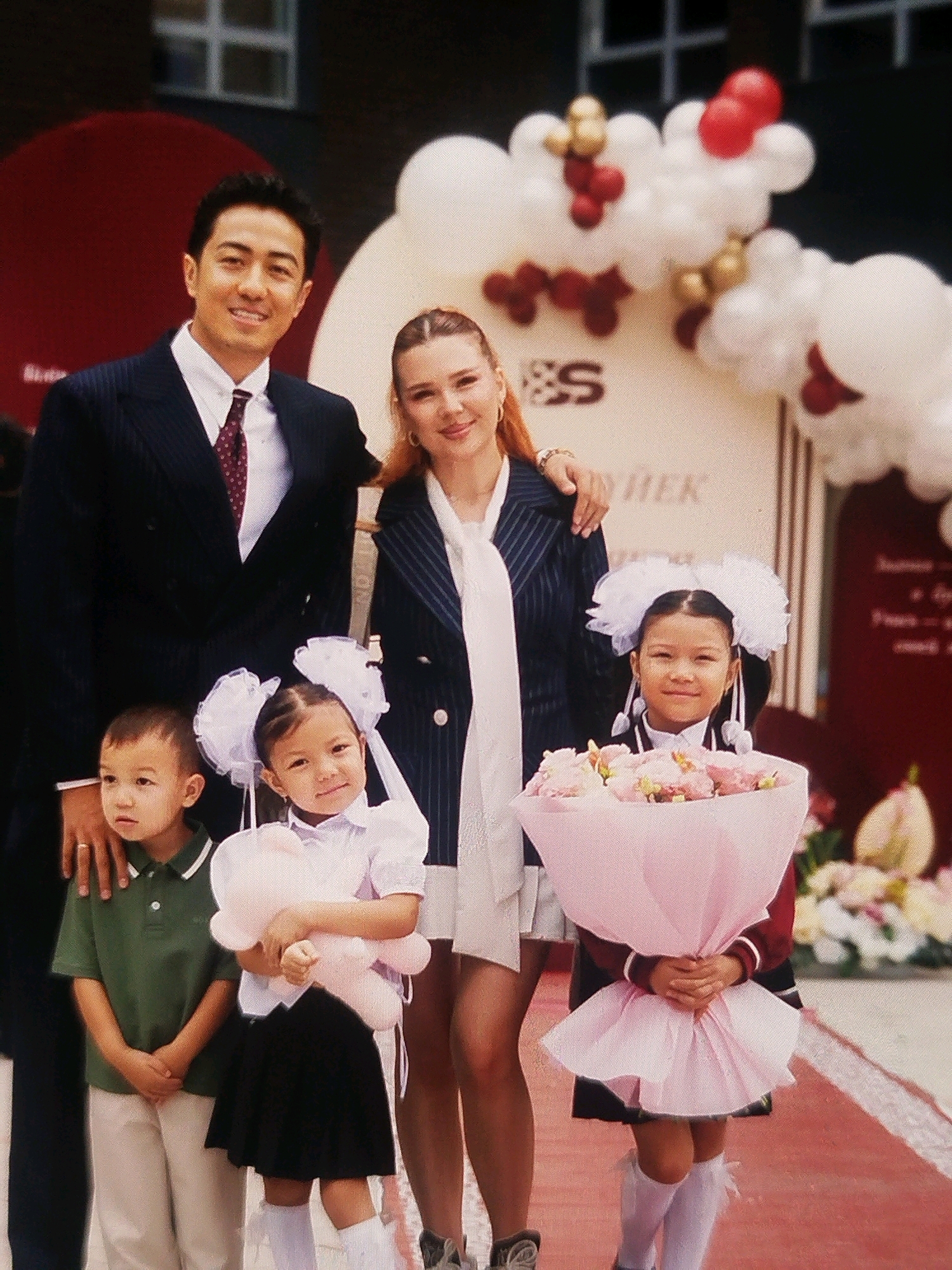 A group of five people, two adults and three children, are posing for a photograph outdoors in front of a building decorated with balloons. The adult male, on the left, is wearing a dark suit with a white shirt and a patterned tie, and he is smiling. The adult female, standing next to him, is wearing a dark blazer over a white blouse with a large white bow tie, and she has her arm around the male.
In the front row, there are three children. On the far left, a young boy in a green polo shirt and tan pants stands with his hands clasped. Next to him, a young girl in a white blouse and black skirt wears large white hair bows and holds a light pink stuffed animal. To her right, another young girl, also in a white blouse and dark skirt, wears large white hair bows and holds a large bouquet of pink and yellow flowers wrapped in pink paper. Both girls are wearing white knee-high socks.
The background features a building with windows and a dark red circular decoration. A festive balloon arch, composed of white, red, and gold balloons, is visible behind the adult female. The ground in front of the group appears to be a reddish carpet or pathway.