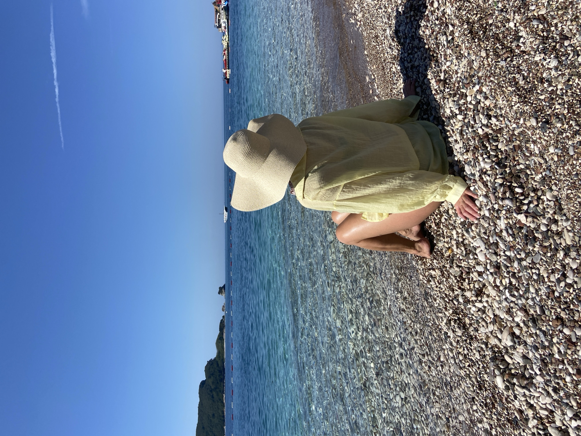 This image depicts an individual seated on a pebble beach, facing a clear blue sea under a cloudless sky. The scene is consistent with coastal characteristics found in the city of Budva, Montenegro.
The subject is an adult female, positioned centrally and viewed from behind. She is wearing a wide-brimmed, light-beige straw hat and a translucent, light-yellow, long-sleeved cover-up. She sits on the beach with her bare legs extended towards the water, her bare feet at the water's edge, and her hands resting on the surrounding pebbles.
The beach surface is composed of smooth, variegated pebbles ranging in color from white and grey to brown and beige. The water at the shoreline is exceptionally clear, allowing visibility of the submerged pebbles, and transitions to a deeper blue further from the shore. Small ripples are visible on the water's surface.
In the mid-distance, a line of red and white demarcation buoys stretches horizontally across the water. On the left side of the horizon, a prominent, dark green, forested landmass, consistent with coastal islands or peninsulas in the Budva area, is visible. To its right, two smaller, rocky islets punctuate the seascape.
Several vessels are present on the water: a white motor yacht is visible to the far left, beyond the buoys; multiple smaller boats are discernible on the far right; and a larger, flat-decked industrial vessel or platform, featuring yellow and white components, is situated in the mid-right portion of the water.
The sky is a uniform, bright blue, with one faint white contrail visible in the upper right quadrant. Direct sunlight illuminates the scene, evidenced by strong shadows cast on the pebbles to the right of the seated individual.