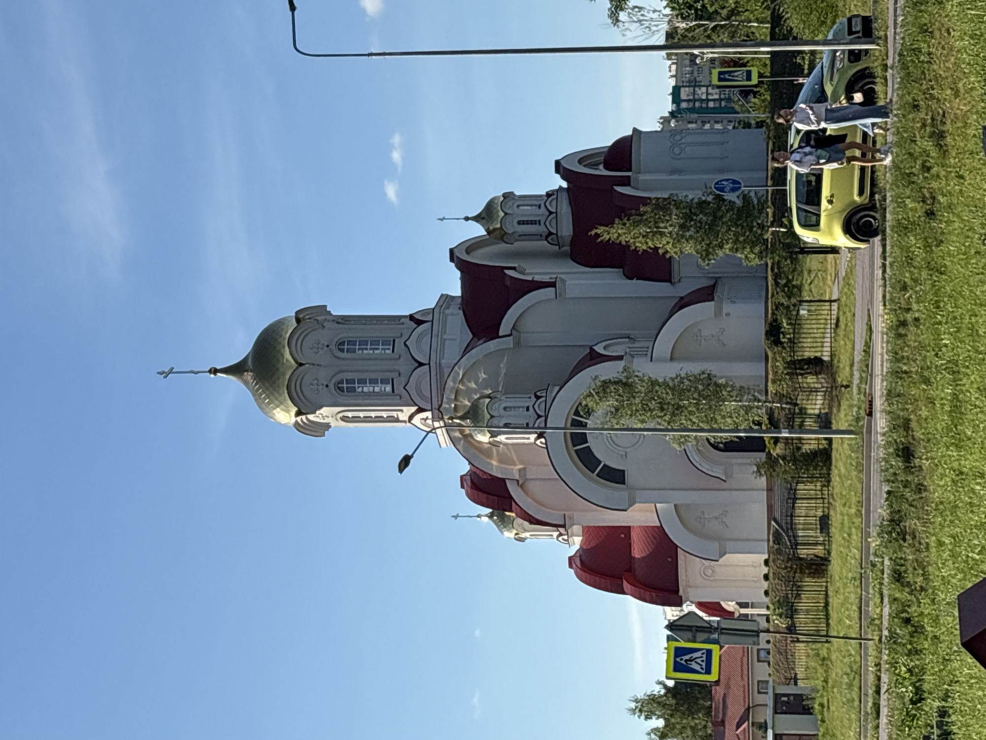 This image captures a street scene in Kaliningrad, Russia, bathed in bright daylight under a clear blue sky with a few wispy clouds. The primary subject is a large, multi-domed building resembling an Orthodox church, positioned on the left side of the frame. The church features white walls, several distinctive red-tiled roofs, and ornate architectural details including arched windows and decorative patterns. Multiple domes, some green and others golden, are topped with Orthodox crosses.
Adjacent to the church, on the right, is a paved street with a grassy verge. Two cars are visible on the road: a prominent lime-green compact car in the mid-ground and another dark green vehicle further down the street. Two individuals are standing near the lime-green car; one is leaning against the vehicle's side, while the other stands close by, suggesting they might be waiting or conversing.
The setting includes a manicured grassy area alongside the street, bordered by a black metal fence, and various green trees providing natural elements. A tall streetlight pole extends into the sky. Two road signs are visible: a square blue sign with a white triangle and a black pedestrian symbol, indicating a pedestrian crossing, and a circular blue sign with a white right-pointing arrow, likely signifying a mandatory direction or turn. The scene conveys a calm, sunny day in an urban environment combining religious architecture with daily street life.