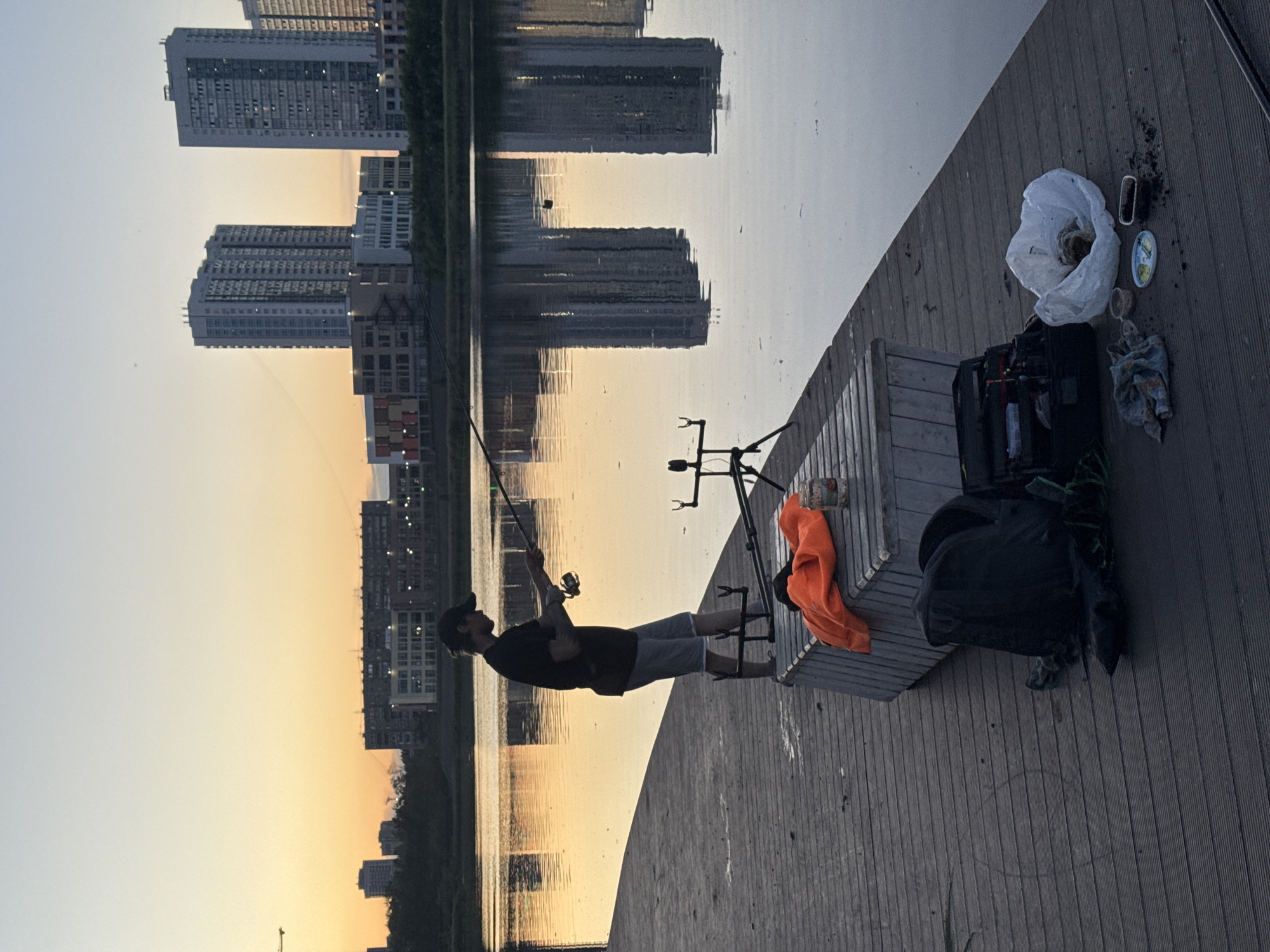 A lone individual is depicted fishing on a wooden pier at dusk in Yekaterinburg, Russia. The scene is set against a backdrop of a serene lake reflecting the warm hues of the setting sun and the silhouettes of distant urban buildings. The person, dressed in a dark t-shirt and shorts, stands with their back to the viewer, engaged in casting their fishing rod. Scattered on the pier are fishing equipment, including rod rests, a tackle box, a backpack, and a bright orange garment placed on a wooden bench. The water is calm, with a few scattered particles on its surface, and the reflections of the skyscrapers on the far side of the lake are clearly visible. The sky transitions from a soft yellow and orange near the horizon to a pale blue overhead, indicating the end of daylight. The overall atmosphere is one of peaceful solitude and twilight recreation.