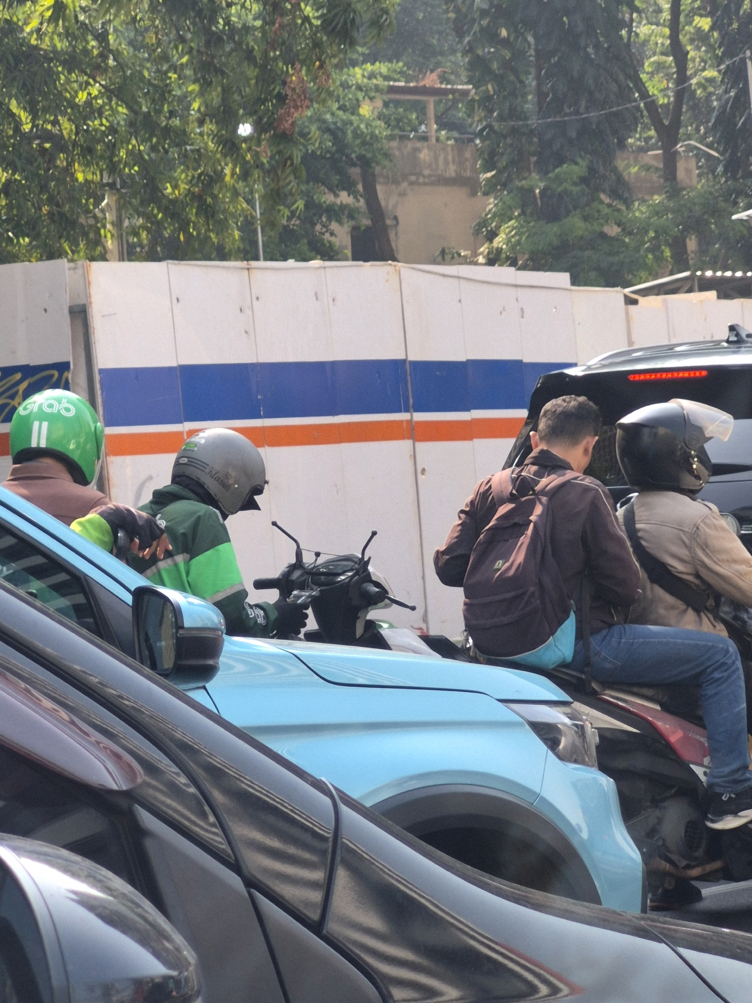 Two motorcycle riders are stopped in traffic on a sunny day in Central Jakarta, Indonesia. The rider in front wears a green helmet with the "Grab" logo and a brown jacket. The rider behind them wears a grey helmet and a green jacket with reflective stripes. They are both on a light blue car, stopped behind several other vehicles. The background shows a white wall with blue and orange stripes and lush green trees.
