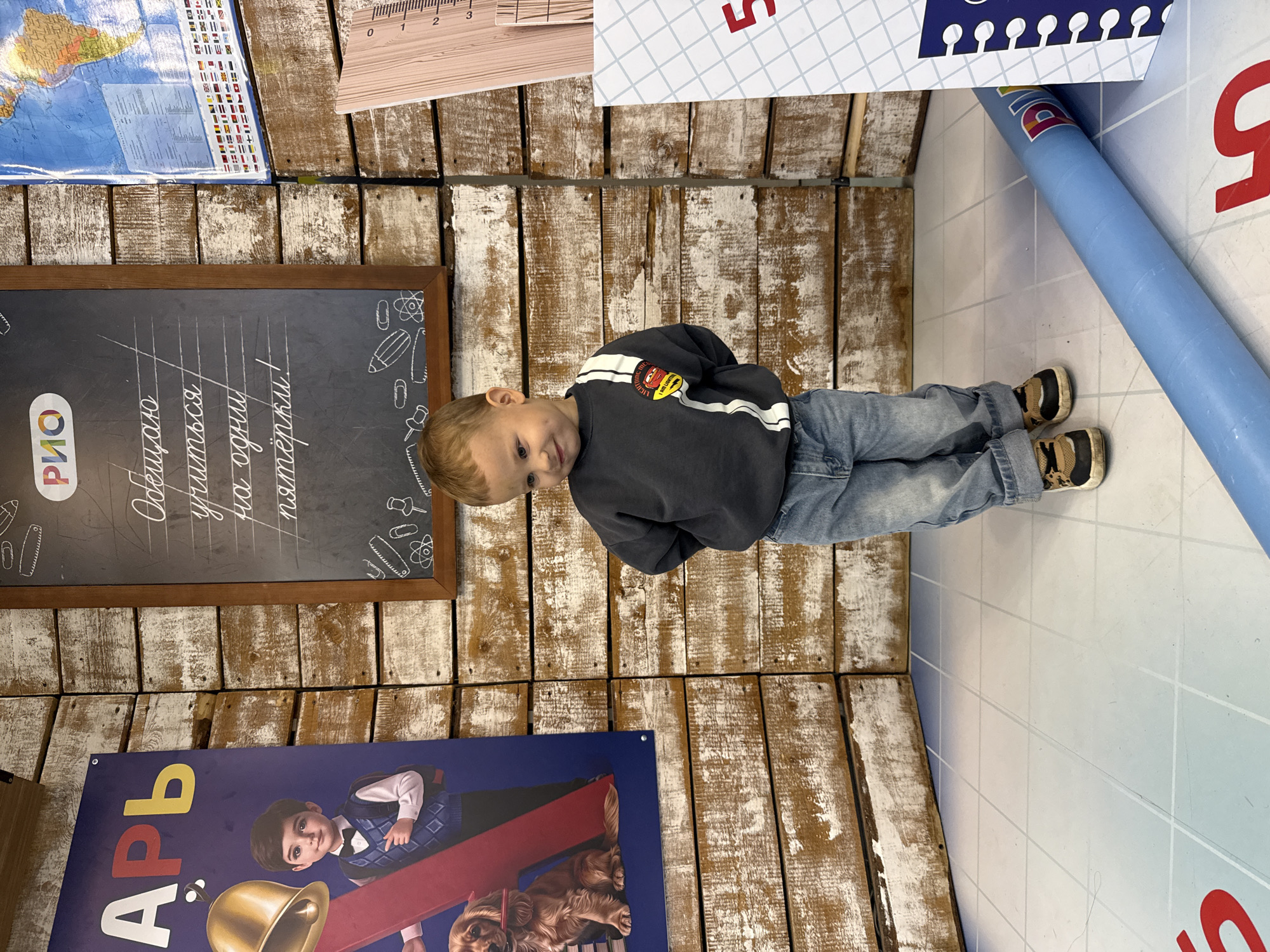 The image depicts a young boy standing in what appears to be a themed indoor space, possibly a children's play area or a photo studio. The background features wooden paneling and decorations reminiscent of a school theme. A large poster with a boy and a dog is visible on the left, and a chalkboard with handwritten Russian text and drawings of school supplies is on the right. A map of the world is also on the wall. The boy is wearing a dark grey sweatshirt with white stripes and a cartoon patch, and blue jeans. He is looking down and to his right with a slight smile. In front of him, a large blue cylindrical object is partially visible, and part of a floor mat with grid lines and red markings is also in view. The lighting suggests an indoor setting. The mention of Korobovo, Russia, as the location is not evident from the visual information in the image itself.