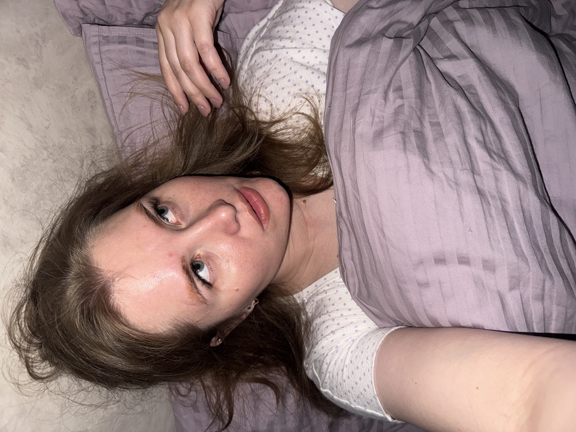 A person is depicted lying on a bed, viewed from a slightly elevated, overhead perspective. The image and crops indicate an indoor setting, likely a bedroom in Gornyi (Dvurechenskoe S/P), Russia, with dim lighting suggesting evening or night.
The main subject is a young person, likely female, with long, light brown hair spread out on a light purple, striped pillow. They are wearing a white top with small purple polka dots. A purple, striped blanket or duvet covers part of their body. Their left hand is visible near their head, showing fingernails with a light, natural-toned polish.
The person's face is prominent, turned towards the viewer. They have fair skin, blue or grey eyes, and a somewhat pensive or weary expression, with their lips slightly parted. Visible details include slight redness on the forehead and nose, and small, metallic hoop earrings.
The activity taking place is simply the person resting or lying in bed. The overall mood is quiet and intimate, emphasized by the soft lighting and the person's relaxed posture. No visible text is present in the media.