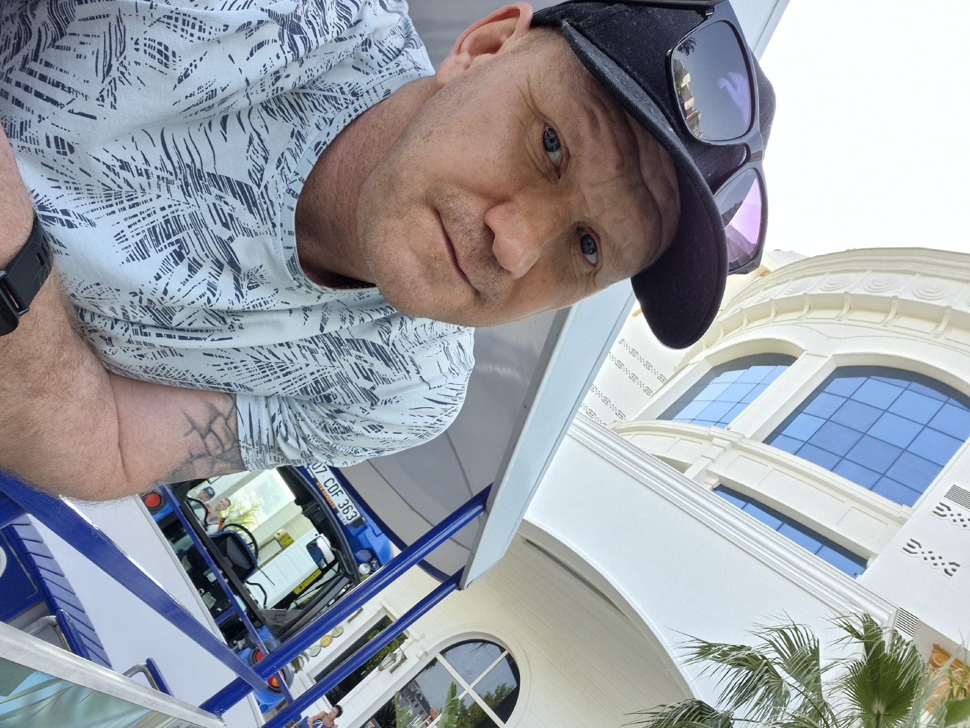 A low-angle, full-color photograph depicts a man wearing a light blue shirt with a dark pattern, a black baseball cap, and sunglasses perched on the cap's brim. He has blue eyes and fair skin. A portion of a black wristwatch is visible on his left wrist, with a tattoo visible on his left forearm. The man is positioned near a blue vehicle with "07 CDF 363" license plate, and the reflection in the vehicle's window shows another person. To the right of the man and slightly behind, is a white building with arched windows and decorative details. A palm tree is visible near the building. The photograph appears to be taken in Alanya, Turkey, suggested by the license plate format of the vehicle.