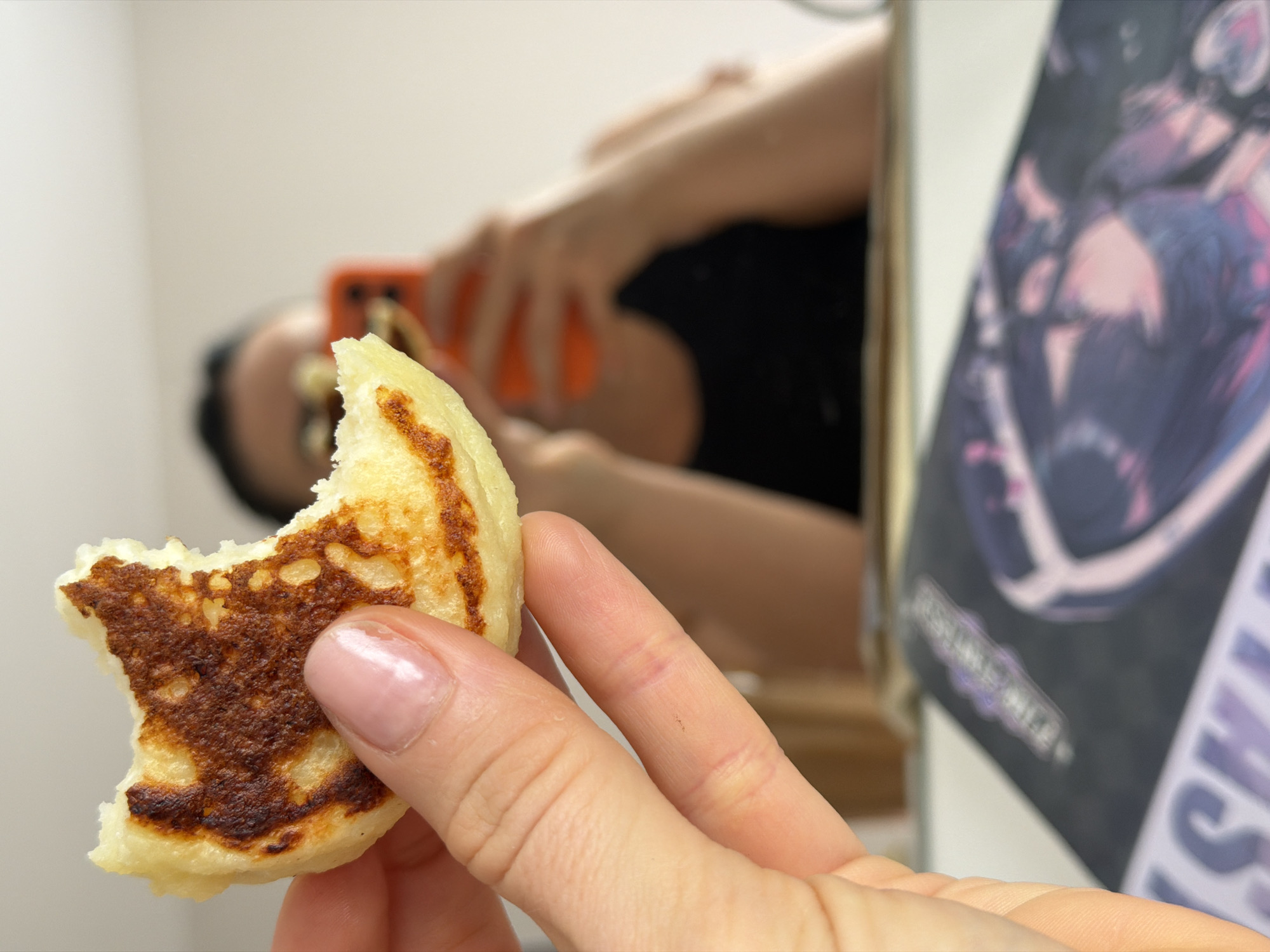 A close-up shot shows a partially eaten syrniki, a fried cheese pancake, held in a person's hand with a well-manicured fingernail. The pancake has a golden-brown crust and a paler interior. In the blurred background, a person is holding a smartphone, potentially taking a photo of the syrniki. Adjacent to the person is a poster with illegible text. The scene appears to be indoors. Given the subject matter and the food item in question, a possible location is Saint Petersburg, Russia, known for its syrniki.