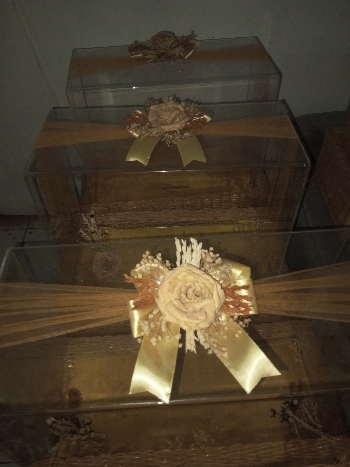 The image displays three clear, rectangular boxes, likely made of acrylic or plastic, stacked vertically against a dark, indistinct background. Each box is intricately decorated with a golden-brown sheer fabric band wrapped around its center. A prominent embellishment adorns the front of each box, consisting of a large, dried, light beige or cream-colored rose-like flower, surrounded by smaller dried floral accents and delicate dried wisps, all topped with a lustrous golden satin bow.
The contents within the transparent boxes are partially visible, appearing to be items made of a woven material, possibly wicker or rattan, suggesting baskets or trays in a natural brown hue. The consistent decorative theme across all three boxes implies they are part of a coordinated set, likely intended for presenting gifts, hampers, or trousseau items for special occasions such as weddings or engagements.
The lighting in the photograph is dim, casting shadows and giving the scene a soft, somewhat muted appearance, characteristic of an indoor setting, possibly during the evening or in a room with subdued artificial light. No people are visible, and no specific activity is taking place; the objects are static, set up for display or readiness. There is no visible text in the image. The overall aesthetic is elegant and celebratory.