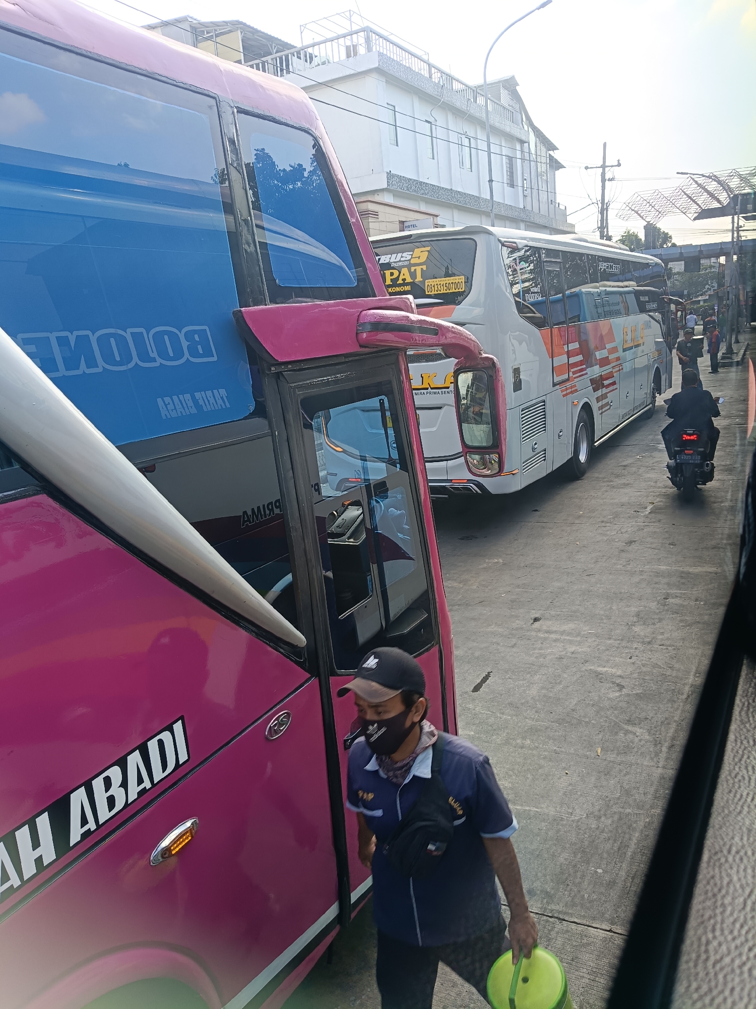 The image shows a street scene in Kedungrejo, Indonesia, with several buses parked and people present. In the foreground, a pink bus is visible, with the text "AH ABADI" on its side. A man wearing a blue uniform, a black face mask, and a black baseball cap is walking towards the right of the frame, holding a green circular object. Behind the pink bus, a white bus with orange and white stripes is parked. Further in the background, another white bus with "BUS 5 PAT" written on it is visible. A motorcycle is also seen on the right side of the image, with its taillight illuminated. The environment appears to be an outdoor public space, possibly a bus terminal or a busy street, with buildings and overhead structures in the background. The lighting suggests it is daytime.