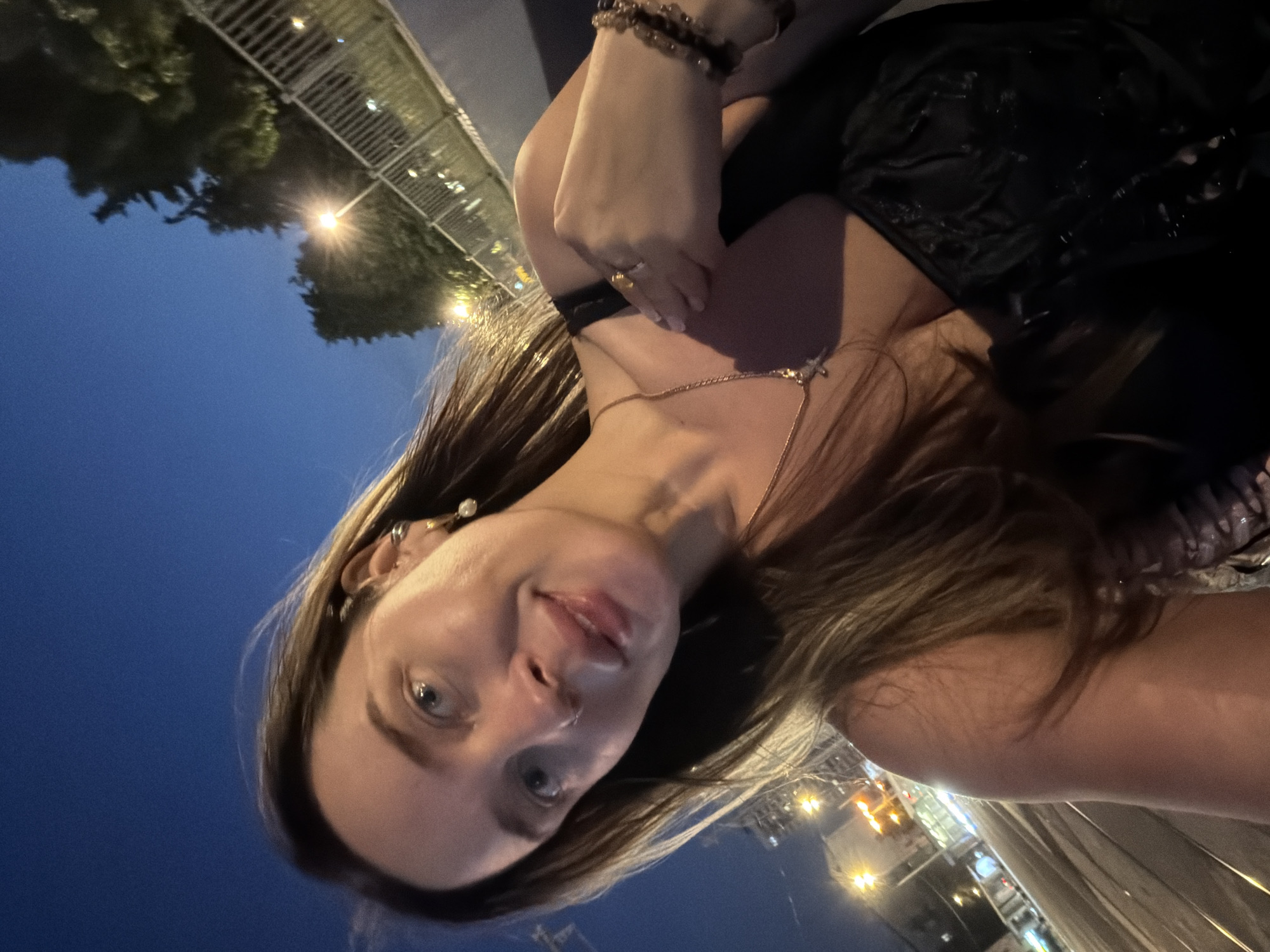 A low-angle, nighttime selfie captures a woman on a city street in Moscow, Russia. The woman, appearing to be in her 20s or 30s, has long, dark brown hair and light blue eyes. She exhibits a subtle smile, looking directly into the camera. She is attired in a dark, possibly black, strapless top featuring textured detailing. Her accessories include a gold-colored necklace with a small pendant, a gold-colored ring on her right hand's ring finger, a multi-beaded bracelet on her right wrist, a pearl drop earring and a smaller stud in her right ear, and a subtle nose stud on her right nostril. The background depicts a dark blue twilight sky over an urban setting illuminated by artificial streetlights. In the lower left, tram tracks are visible in the foreground, leading towards distant city lights. To the right, a metal railing runs horizontally, bordering a treeline that is lit by a prominent streetlamp and other distant lights.