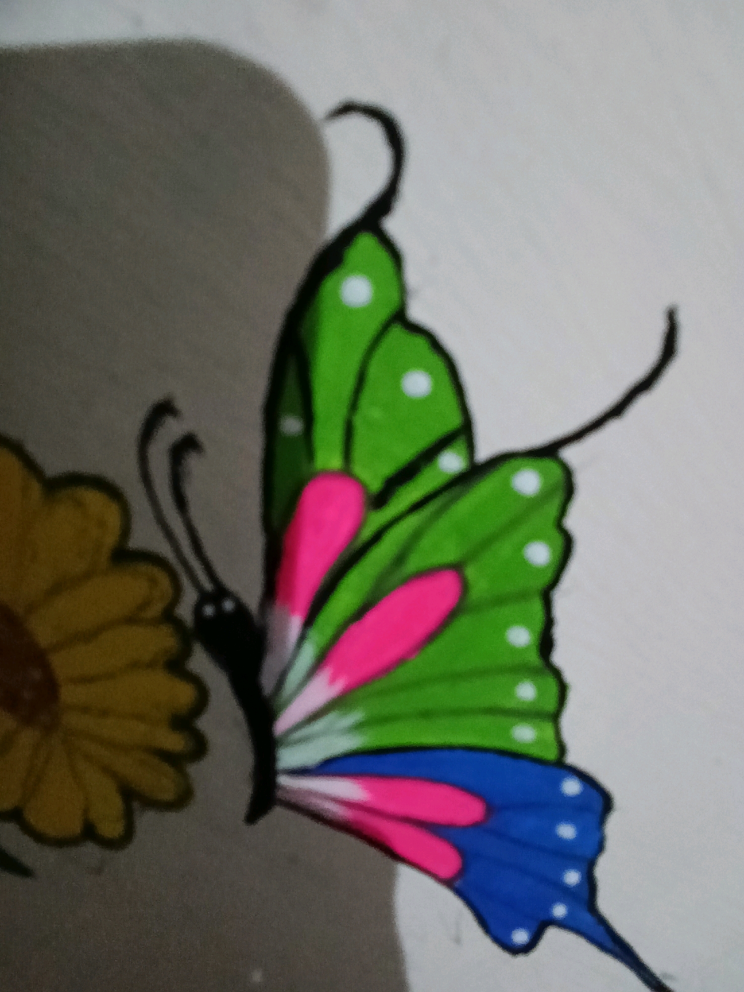 The image shows a drawing of a butterfly with green, pink, and blue wings, adorned with white polka dots. To the left of the butterfly is a drawing of a yellow flower. The drawing appears to be on a light-colored surface, possibly paper or a wall, with a gray shadow cast on the left side. The drawing is not located in Lehra Sondha, India, as there is no information within the image to suggest a geographical location.