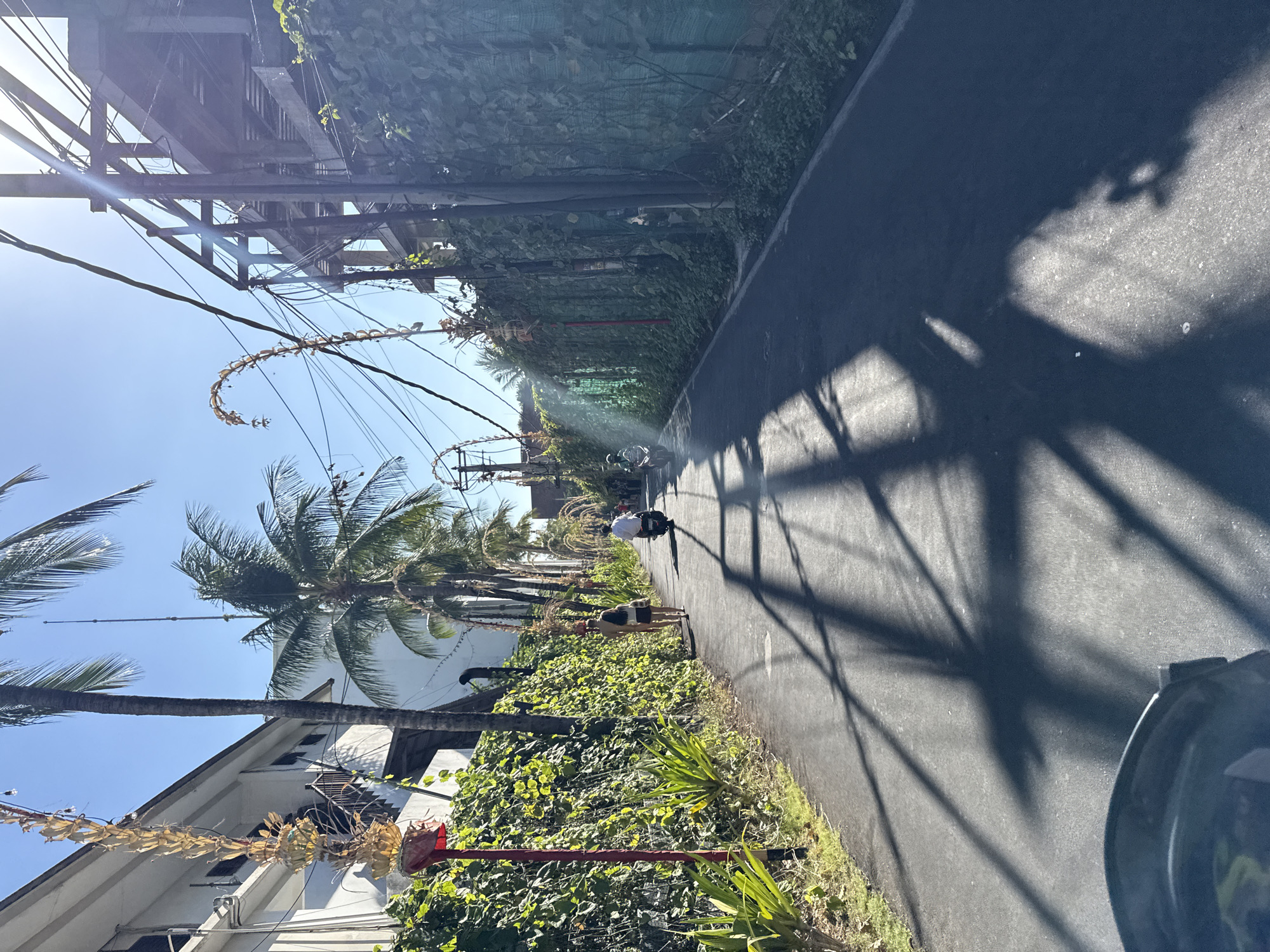 The image depicts a sun-drenched, narrow paved road in North Kuta, Indonesia, lined with lush green foliage and buildings. A couple walks away from the camera on the left side of the road, while a person on a scooter approaches from the distance on the right. Palm trees and overhead utility wires are visible, with the sun casting long, dramatic shadows across the road. The scene appears to be a typical street view in a tropical urban environment.