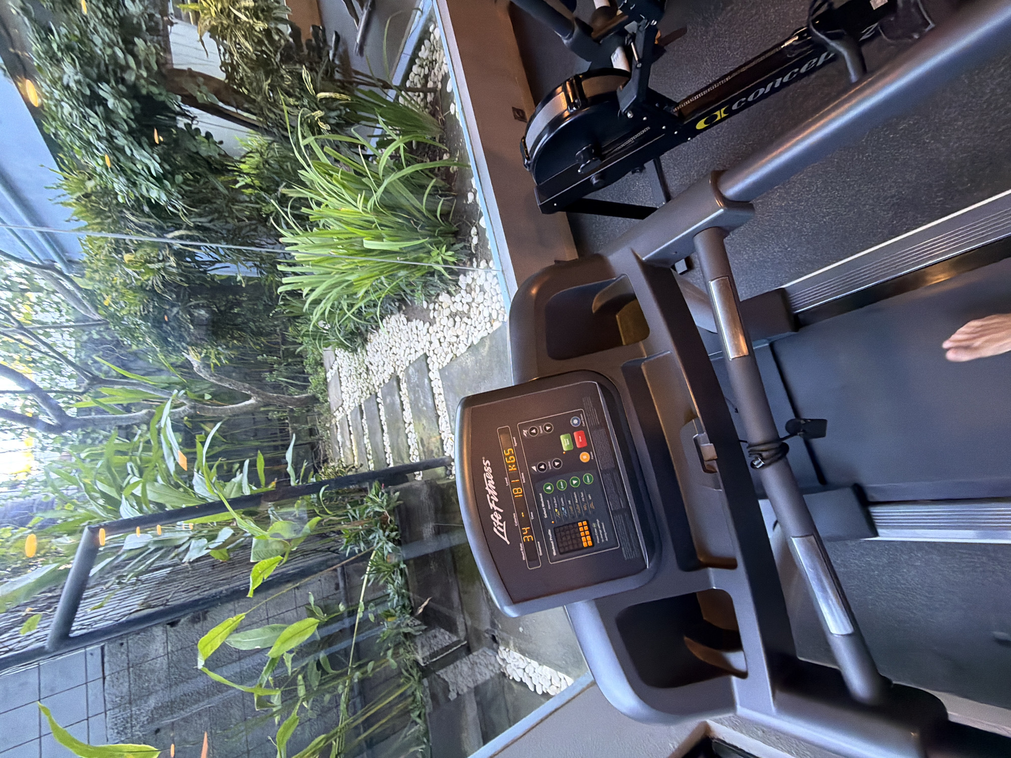 A close-up perspective shows a Life Fitness treadmill, featuring a display indicating a weight of 181 KG, situated in what appears to be a gym setting in Kuta, Indonesia. Adjacent to the treadmill is a Concept2 rowing machine. The setting is characterized by a garden view, with greenery and white gravel visible through a window. A person's foot is slightly visible at the edge of the treadmill belt. The location is identifiable as being in Kuta, Indonesia, based on the typical architecture, landscaping, and gym equipment often found in fitness centers in that area.
