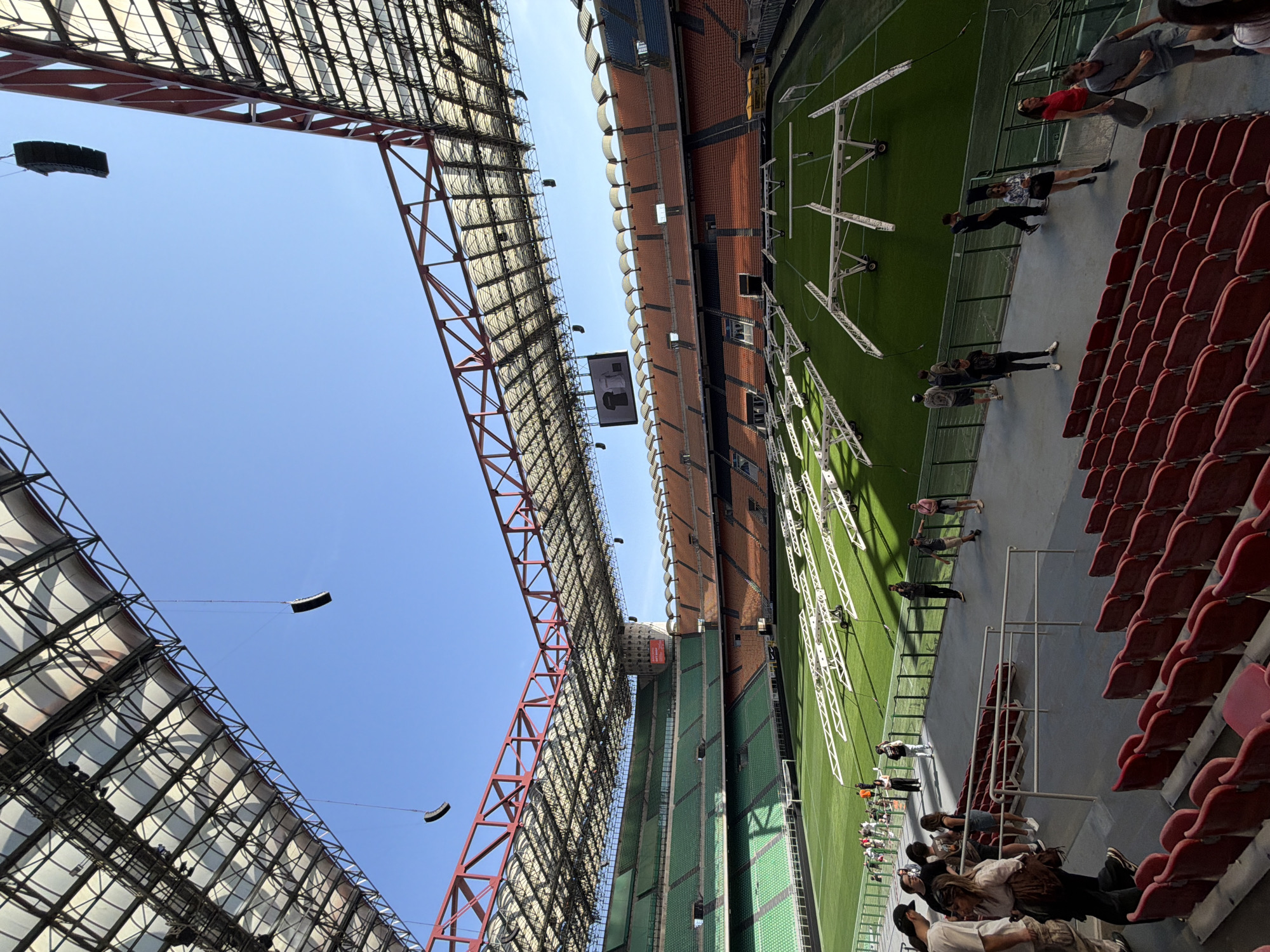 The image shows the Stadio Giuseppe Meazza, also known as San Siro, a stadium in Milan, Italy. The stadium is an open-air stadium with a roof structure visible on both sides. Under the roof, there are suspended dark objects, possibly speakers or lights. The stadium's seating area is visible on the right, with rows of red seats. The playing field is visible, with what appears to be equipment or structures set up on it, perhaps for an event. Several people are visible on the field and near the seating area, suggesting ongoing activity or preparation. The sky is clear and blue.