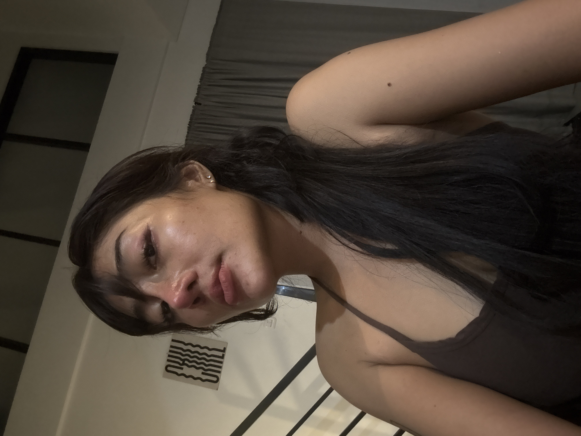 A close-up, slightly angled photograph depicts a woman with long, dark hair wearing a camisole-style top. Her skin is visible, and she has a mole on her upper arm. An earring is visible in her left ear. The background includes a window and curtain. A sticker is on the wall. A banister for stairs is visible.