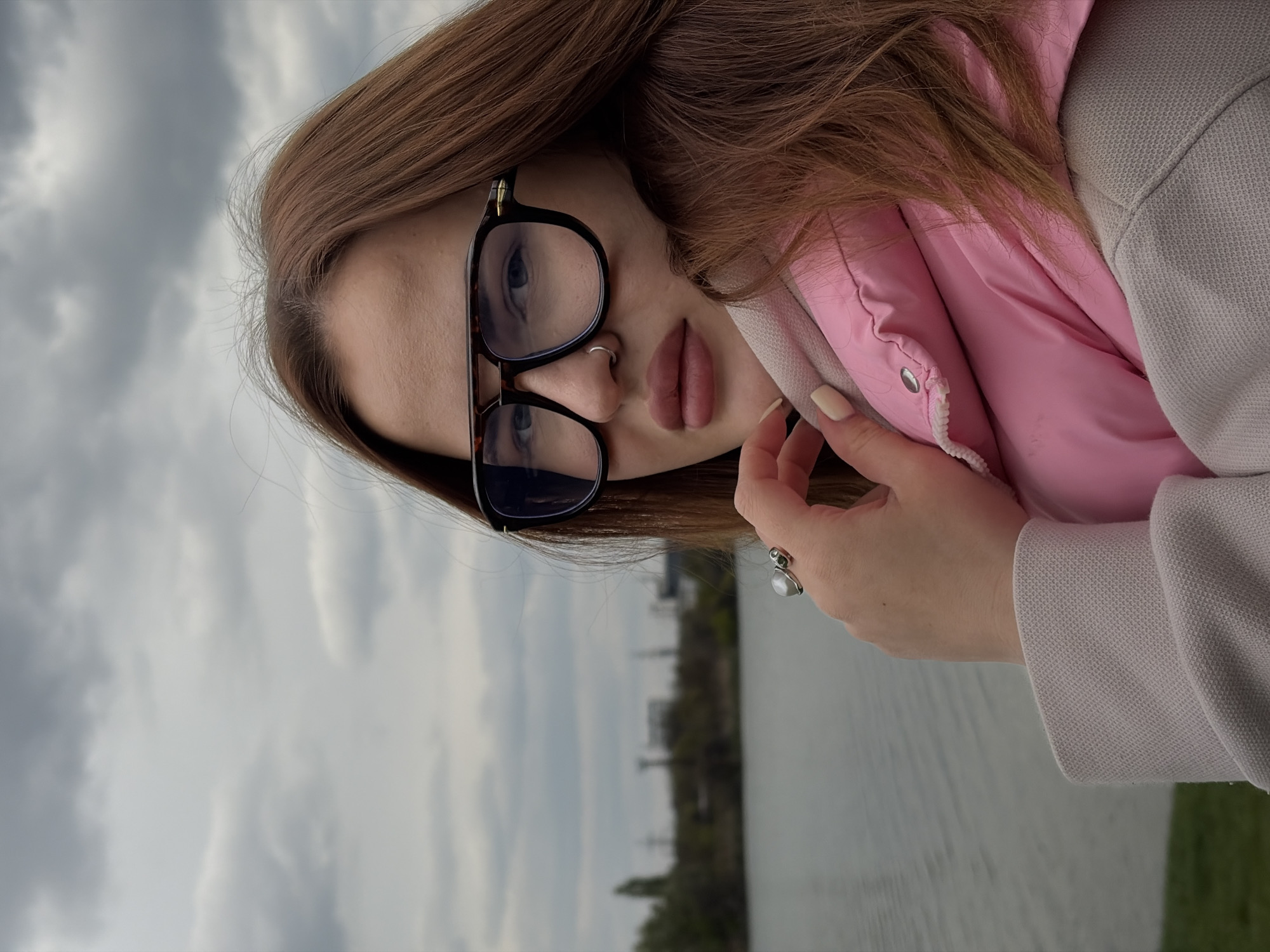 A young woman with long brown hair and black-rimmed glasses is posing outdoors. She is wearing a pink puffer jacket under a beige coat. She has a ring on her finger and a nose ring. In the background is a cloudy sky, water, trees, and indistinct buildings, suggesting the scene is in an urban environment. While it's difficult to definitively confirm, based on visual analysis of the environment, it could plausibly be in Krasnodar, Russia.