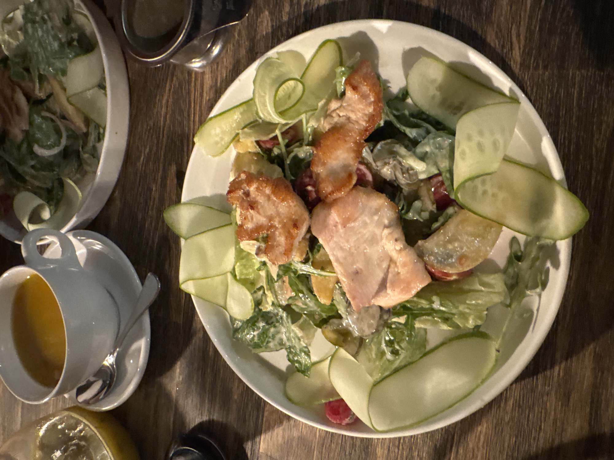 A close-up overhead shot captures a dining table setting with two plates of food and a cup of soup. In the foreground, a white plate holds a salad with grilled chicken pieces, cucumber slices, and leafy greens coated in dressing. To the left, a white cup and saucer contain a yellow-orange liquid, presumably soup, with a silver spoon resting on the saucer. In the background, a second white bowl contains a similar salad with cucumber ribbons and greens. A dark wooden table provides the surface for these items. The scene appears to be indoors, likely in a restaurant, but no specific location within Perm, Russia can be identified from the image.