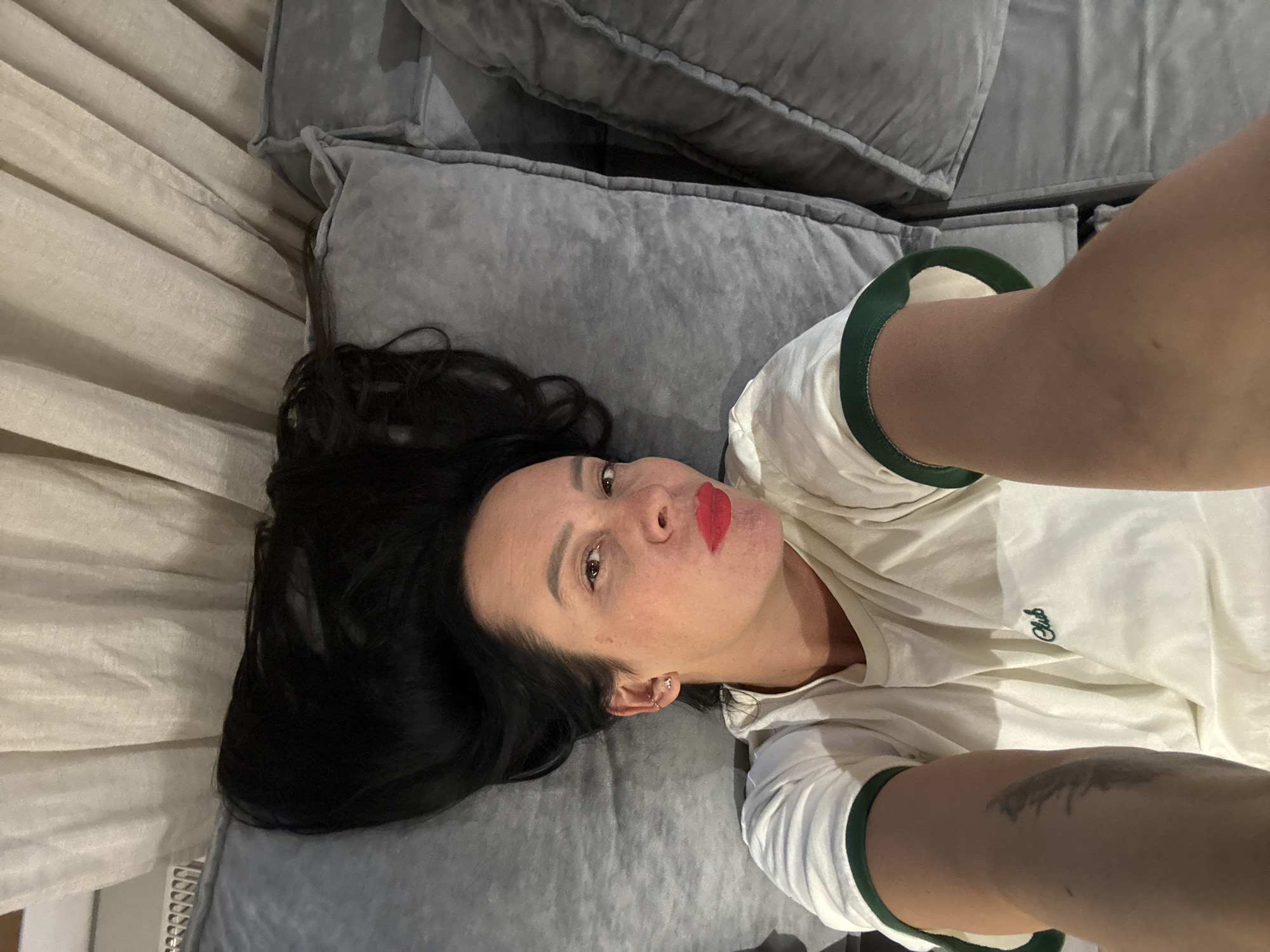 A person is lying on a grey sofa or large cushion. The person has long, black hair spread out on the sofa. The person is wearing a white t-shirt with green trim around the sleeves and neck. The person is wearing bright red lipstick. The person is fair-skinned and has visible light brown or grey eyes. A grey, textured curtain or wall covering is visible to the left of the person's head. A tattoo is visible on the forearm extending towards the camera. The context suggests that the image was taken as a selfie, and the location is presumably indoors. There is no information in the image to confirm the location is Chaikovsky, Russia.