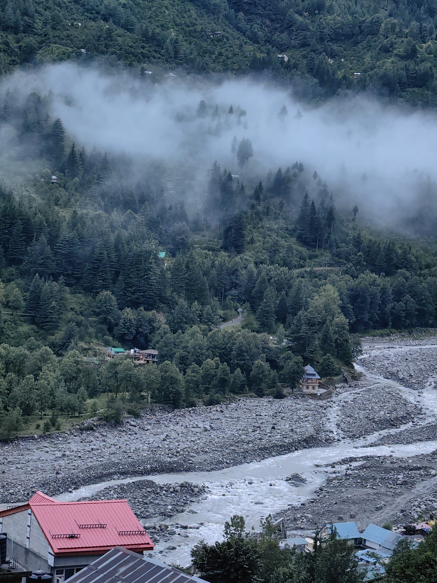 This image captures a misty morning scene in Vashisht, India, with a focus on the natural landscape and scattered human settlements. The foreground features a house with a prominent red, corrugated metal roof and another with a grey metal roof, suggesting a residential area. Adjacent to these structures is a wide, rocky riverbed with a churning, grey river flowing through it.
The midground is dominated by a dense forest of pine trees covering a steep hillside. A winding road is visible cutting through the trees, and a few small buildings with dark roofs are nestled amongst the foliage, indicating scattered homesteads or guesthouses. A small, multi-story building with a brown and tan facade and a dark roof stands near the riverbank, possibly a temple or a notable structure.
The background is a continuation of the forested hillside, shrouded in a thick layer of white mist that partially obscures the upper reaches of the mountains and some distant structures. The mist creates a serene and somewhat mysterious atmosphere, highlighting the natural beauty and isolated settlements of the region. The overall impression is one of a tranquil, mountainous environment with evidence of human habitation integrated into the landscape.