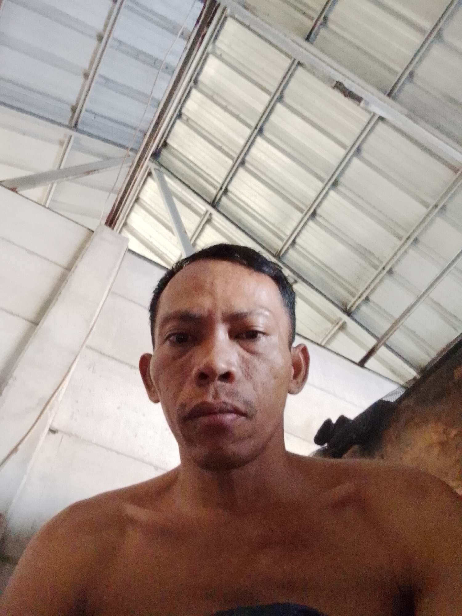 The photograph shows a man with short black hair and brown skin, facing the camera. His upper body is bare. The location is possibly Bukit Ketok, Indonesia, as indicated. The background consists of a white wall and a corrugated metal roof with a supporting steel frame.