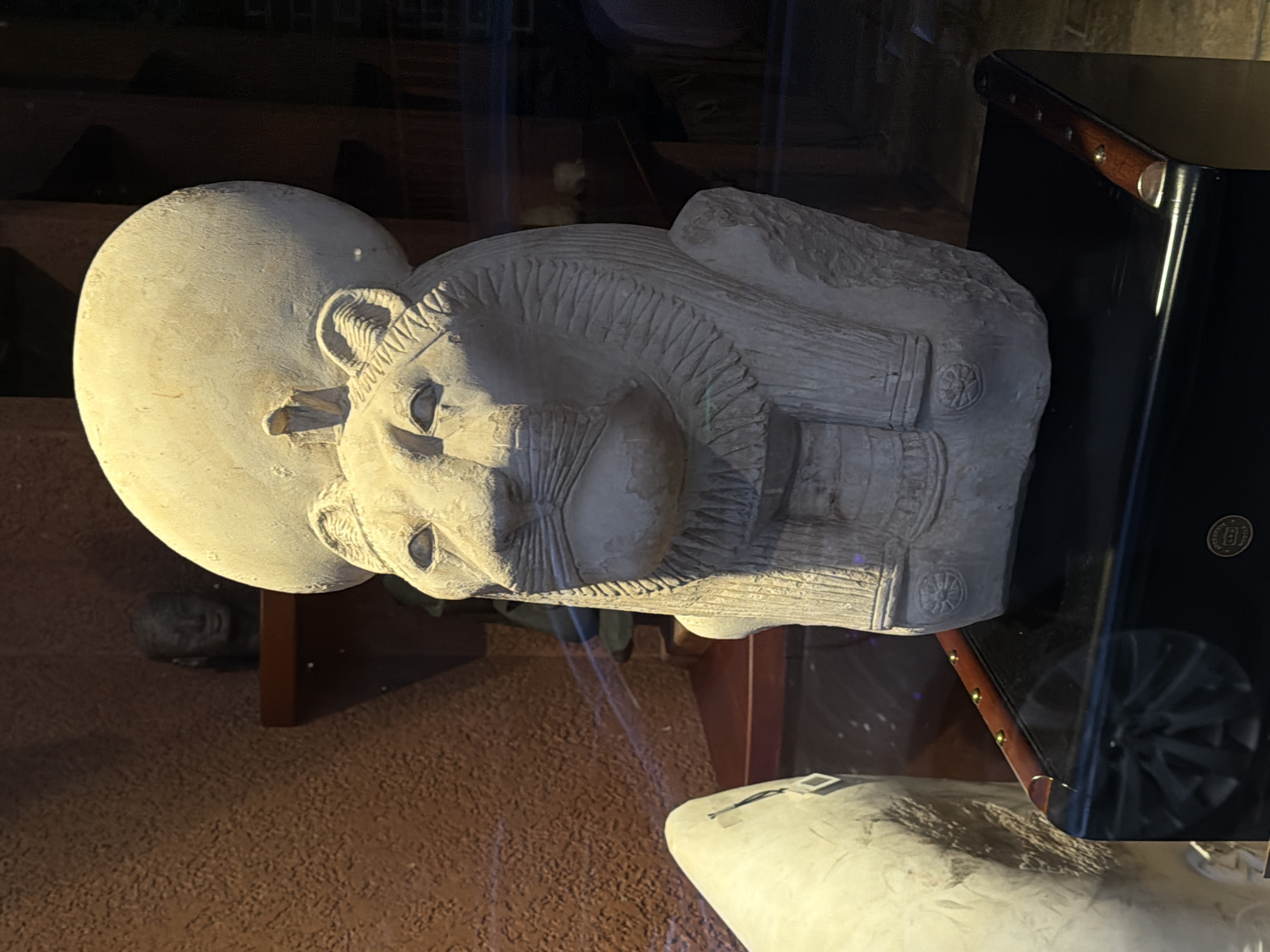 The image depicts a head of Sekhmet in the National Museum of Serbia, Belgrade. The statue is made of stone and has a sun disk atop her head. A cabinet is on the right side. The flooring and shelving are reddish-brown. There appears to be another statue located beneath the object of focus. The image looks to have been taken through a glass barrier of some kind.
