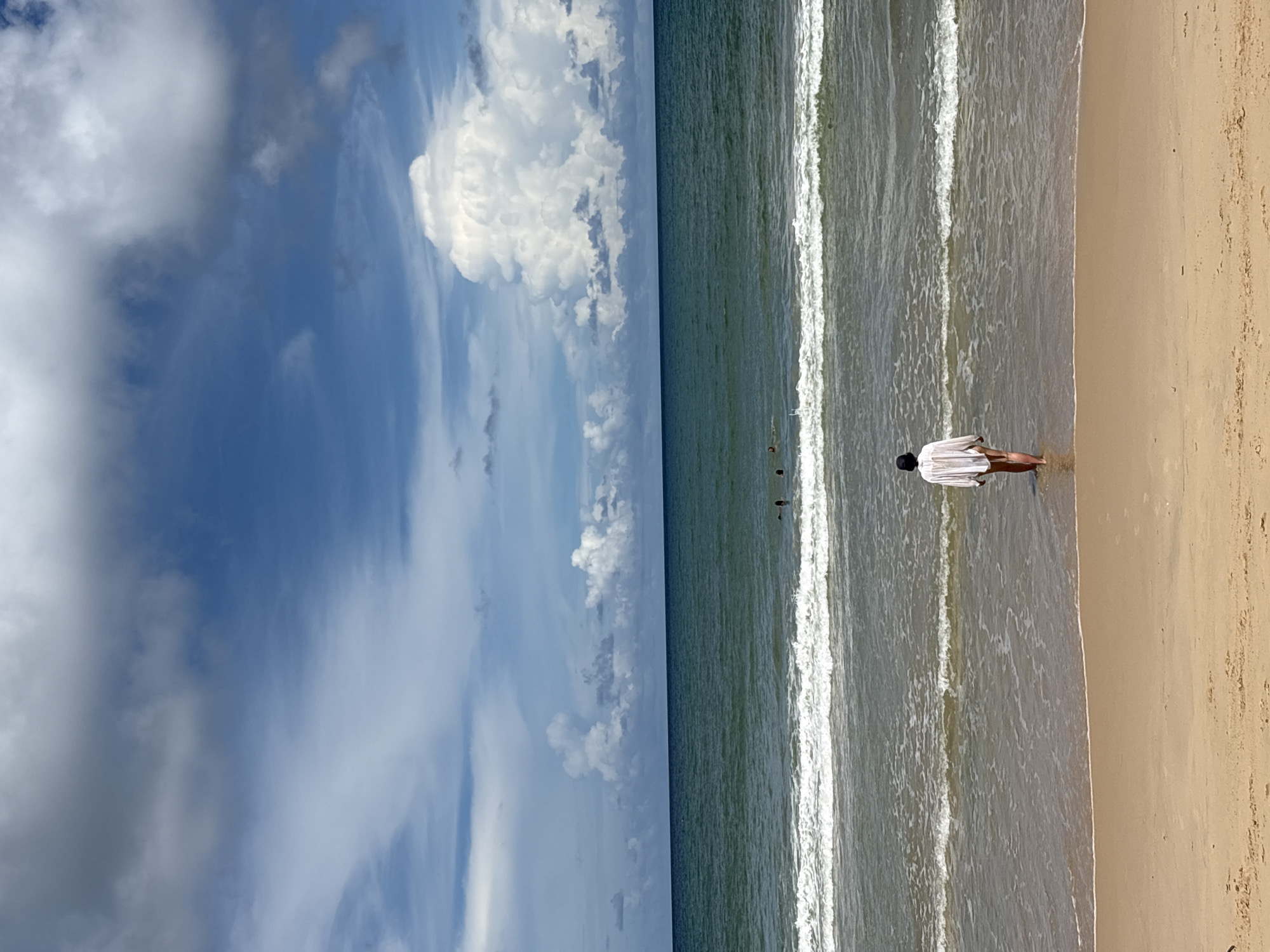In the city of Choeng Thale, Thailand, this image captures a person from behind, wading into the ocean. The individual is wearing a loose white shirt and dark bottoms, with their legs visible as they step into the water. In the distance, several other people are swimming or wading in the sea, and waves are breaking on the shore. The sky above is a mix of blue and white clouds, with a large, dramatic cloud formation dominating the upper right portion of the frame. The scene is set on a sandy beach, with the ocean stretching out to the horizon.