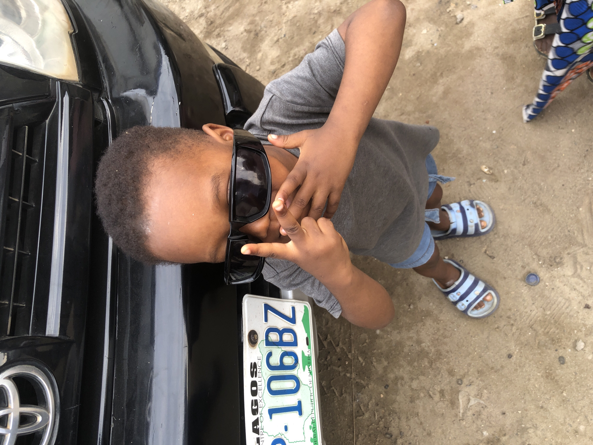 A young child wearing sunglasses and a gray t-shirt stands in front of a black SUV. The child's hands are covering their mouth and nose. The SUV's license plate is visible and reads "LAGOS P-106BZ FEDERAL REPUBLIC OF NIGERIA". The ground is sandy and appears to be in an outdoor setting. The overall scene is casual and appears to be taken in a residential or street area in Ado, Nigeria.