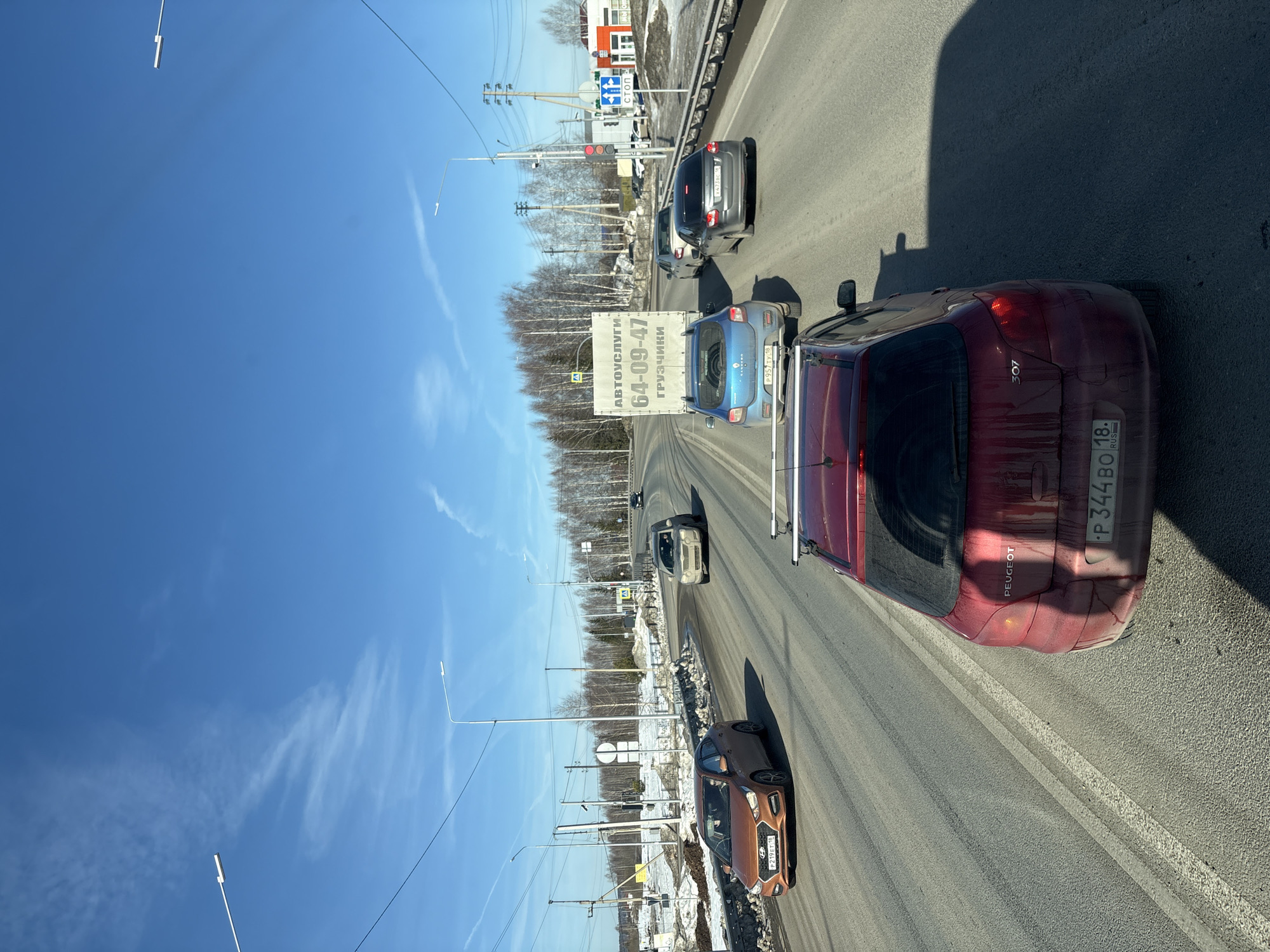 The image depicts a daytime traffic scene on a multi-lane asphalt road in Staroe Mikhailovskoe, Zav'yalovsky District, Udmurt Republic, Russia. The sky is clear blue with sparse white wispy clouds.
In the foreground, the rear of a dirty, dark red Peugeot 307 hatchback (license plate P344BO 18 RUS) is visible, equipped with roof racks. Directly ahead in the same lane is a light blue Dacia/Renault Sandero van (license plate P957TY 18 RUS) featuring a prominent white cargo box on its rear, labeled "АВТОУСЛУГИ 64-09-47 ГРУЗЧИКИ" (Auto Services 64-09-47 Movers). To the left of the van, in an adjacent lane, an orange Hyundai Solaris sedan (license plate P219ET 18 RUS) is driving alongside. Further down the right lane, a dark grey sedan (license plate X473BC 18 RUS) is observed, followed by a light grey SUV. All visible vehicle license plates bear the "18" region code, confirming the Udmurt Republic.
The road surface shows signs of wear and dirt, with clear white lane markings. Piles of melting snow and slush are accumulated along the road shoulders and median, indicative of early spring conditions. The surrounding environment consists of bare deciduous trees, possibly birch, lining the background. Numerous utility poles with overhead lines and modern streetlights with LED fixtures are present along the roadway. On the right side, an active red traffic light is visible, indicating a stop. Alongside it, blue and white road signs display a "СТОП" (STOP) symbol and an arrow indicating a mandatory right turn. A triangular pedestrian crossing sign is also visible. Further down on the right, a commercial building with an orange roof and white walls is situated. No specific individuals are clearly discernible in the vehicles or on the roadside.