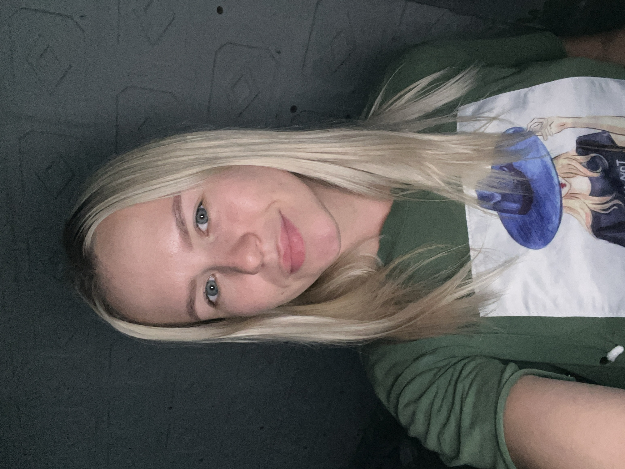A blonde woman with blue eyes and fair skin is pictured in a close-up selfie. She is wearing a green t-shirt with a graphic print of a woman wearing a blue hat. The background appears to be a dark, textured wall. The lighting is dim, casting shadows on her face and creating a moody atmosphere. The woman is looking directly at the camera with a slight smile.