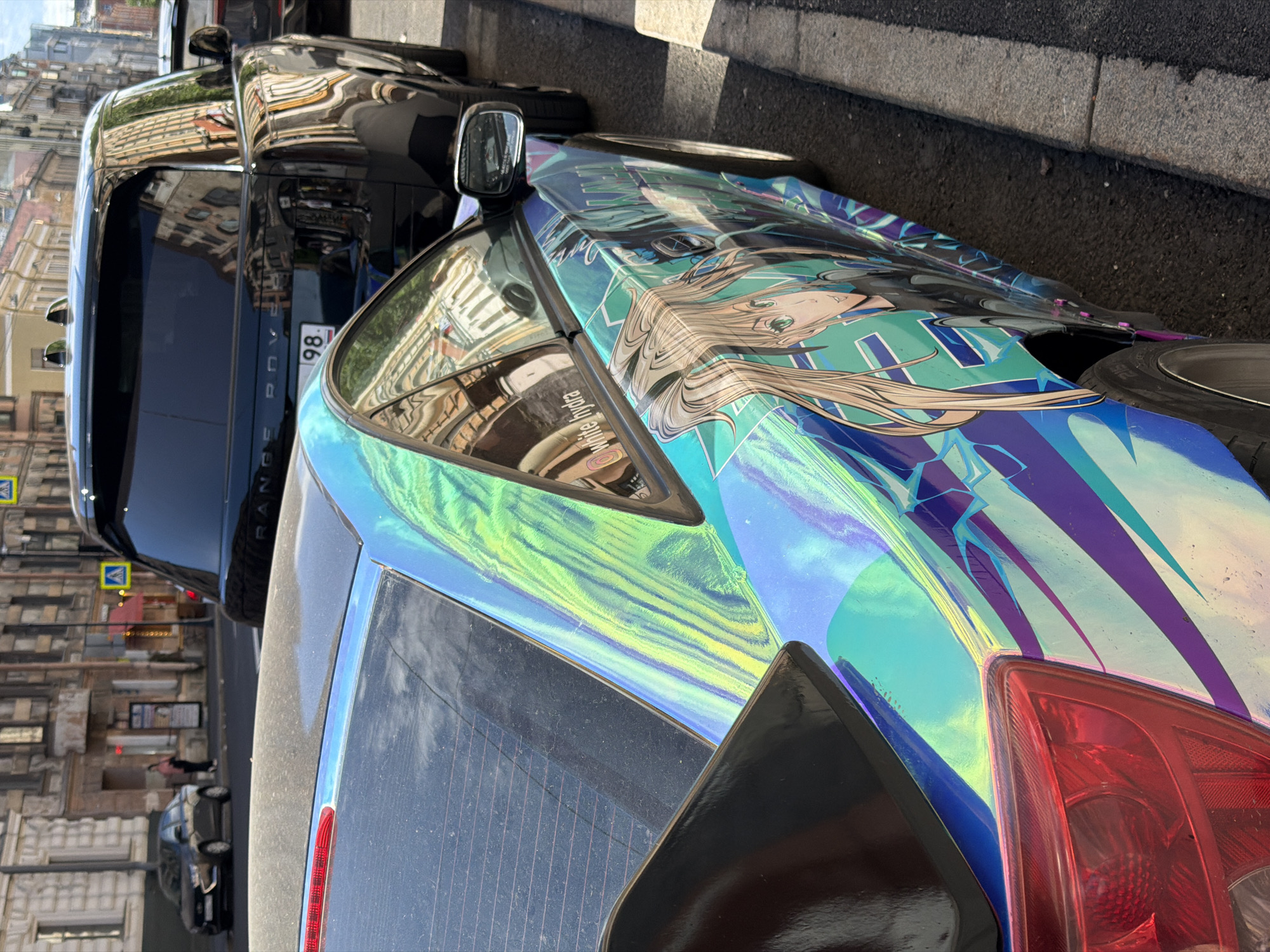 The image shows a close-up, angled view of two parked cars on a street in Saint Petersburg, Russia. The foreground car is a dark-colored vehicle with a striking, iridescent wrap featuring an anime-style illustration of a female character with long blonde hair and green eyes. The wrap has a holographic effect, shifting between shades of green, blue, and purple. The car's rear taillight is visible on the right side, and a black spoiler is present. Behind it, a black Range Rover is parked, with its license plate partially visible, reading "98". The reflection of buildings with architectural details typical of Saint Petersburg can be seen in the Range Rover's windows, suggesting an urban setting. The lighting indicates it is daytime, likely late morning or afternoon, with bright sunlight casting shadows. The overall scene depicts urban street parking with attention drawn to the customized, eye-catching design of the foreground car. No people are clearly visible.