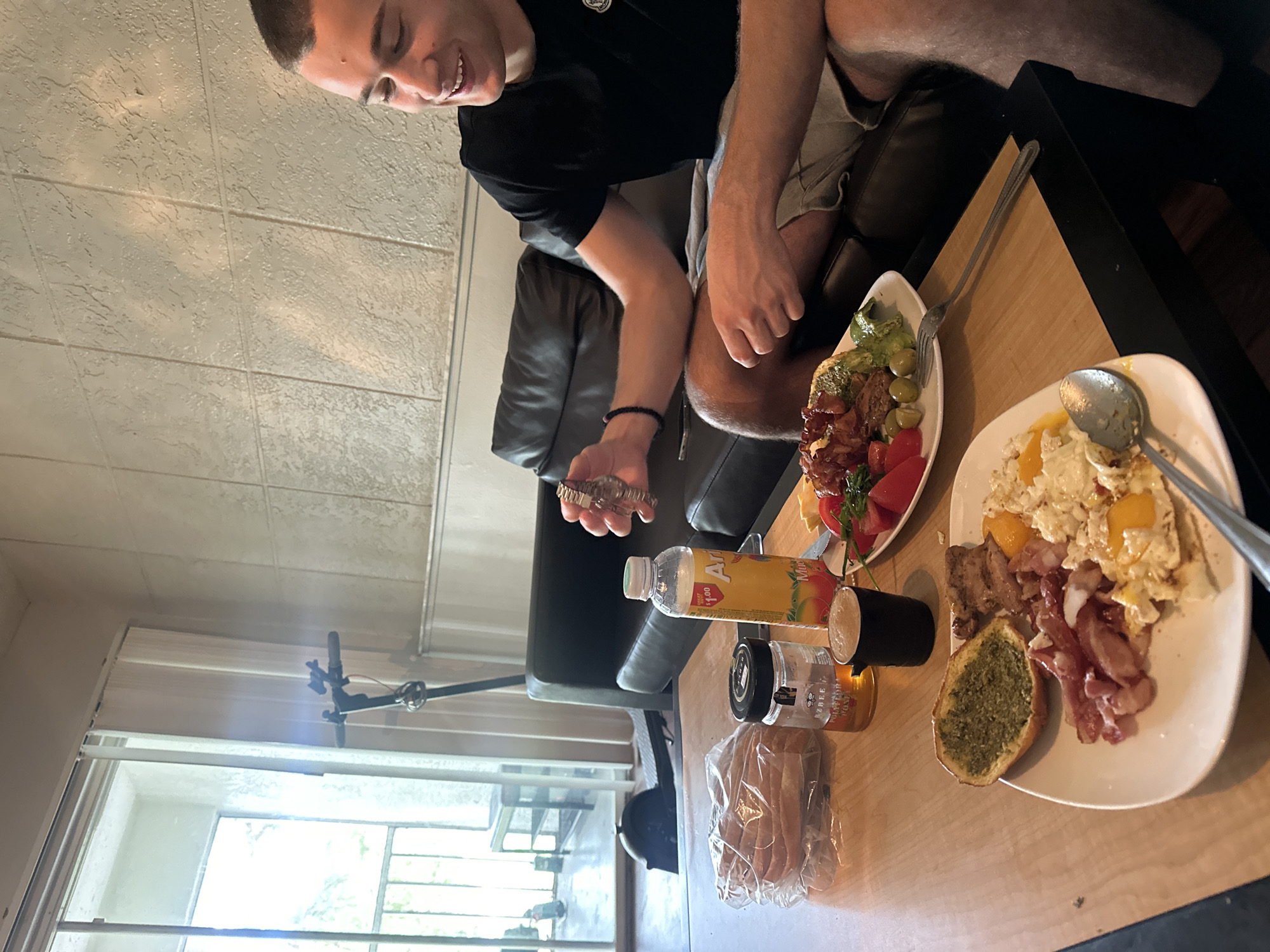 A medium shot depicts a male in a black shirt, seated on a dark leather sofa at a wooden table, presumably indoors. The man is looking down and holding a wristwatch. The table features two plates of food: one with scrambled eggs, bacon, and a slice of garlic bread, and another with bacon, tomatoes, avocado, and olives. A bottle of AriZona iced tea, a jar of honey, and bread in a plastic bag are also on the table. Outside the window, there appears to be a view of an outdoor seating area. The location is University, United States of America.