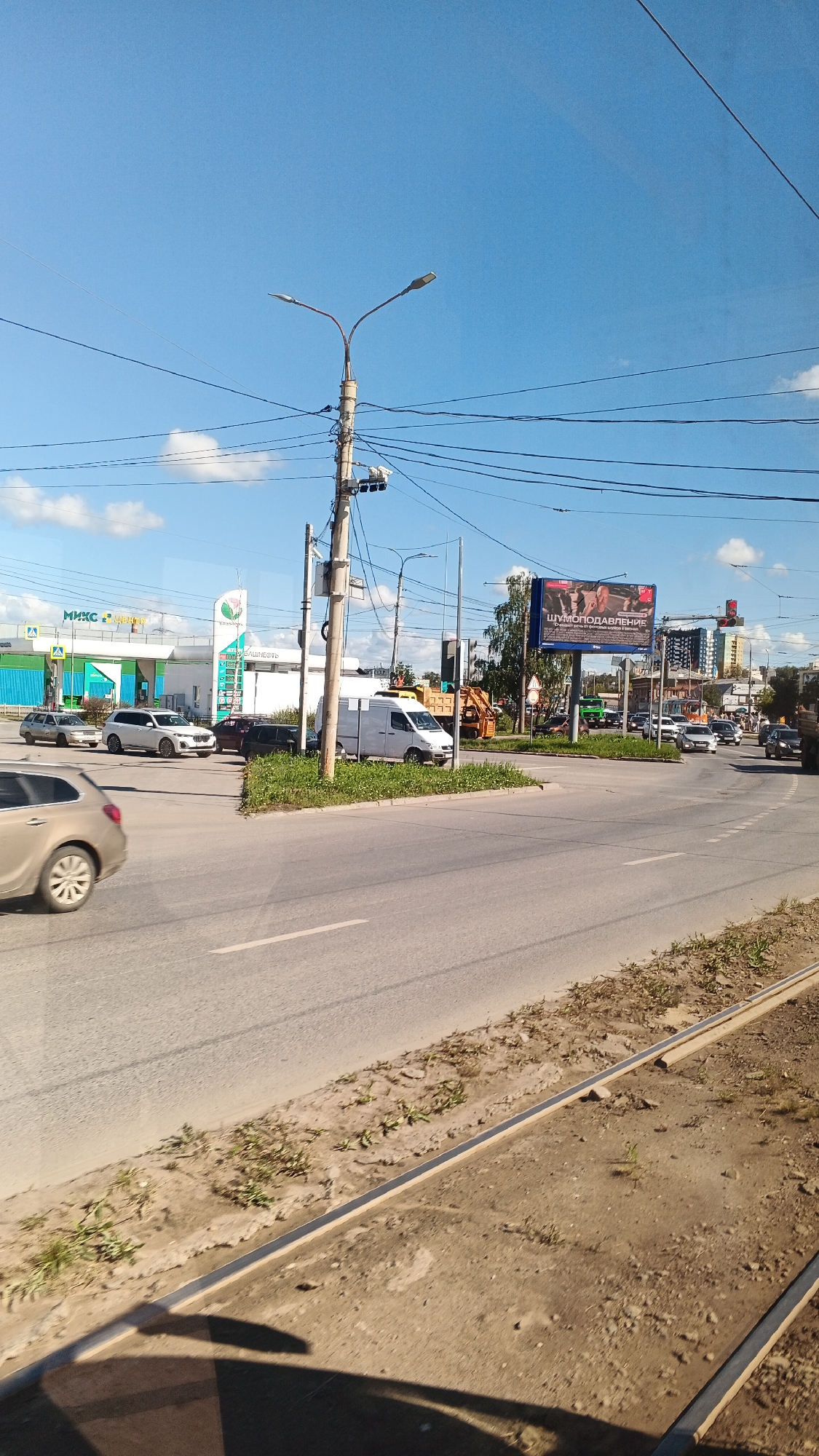 The image depicts a street intersection in Izhevsk, Russia, on a clear day with blue skies and scattered clouds. The foreground shows a grey asphalt road with white lane markings and a dirt shoulder with sparse vegetation. In the mid-ground, several cars are visible, including a light-colored SUV, a dark sedan, and a white van. A traffic light and a street lamp with multiple lights are prominent on the right side of the intersection, attached to a utility pole with numerous wires. A billboard advertising "ШУМОПОДАВЛЕНИЕ" (noise suppression) is visible in the background. Further back, buildings and trees line the street, suggesting an urban or suburban environment. The overall scene is typical of a city street with moderate traffic.