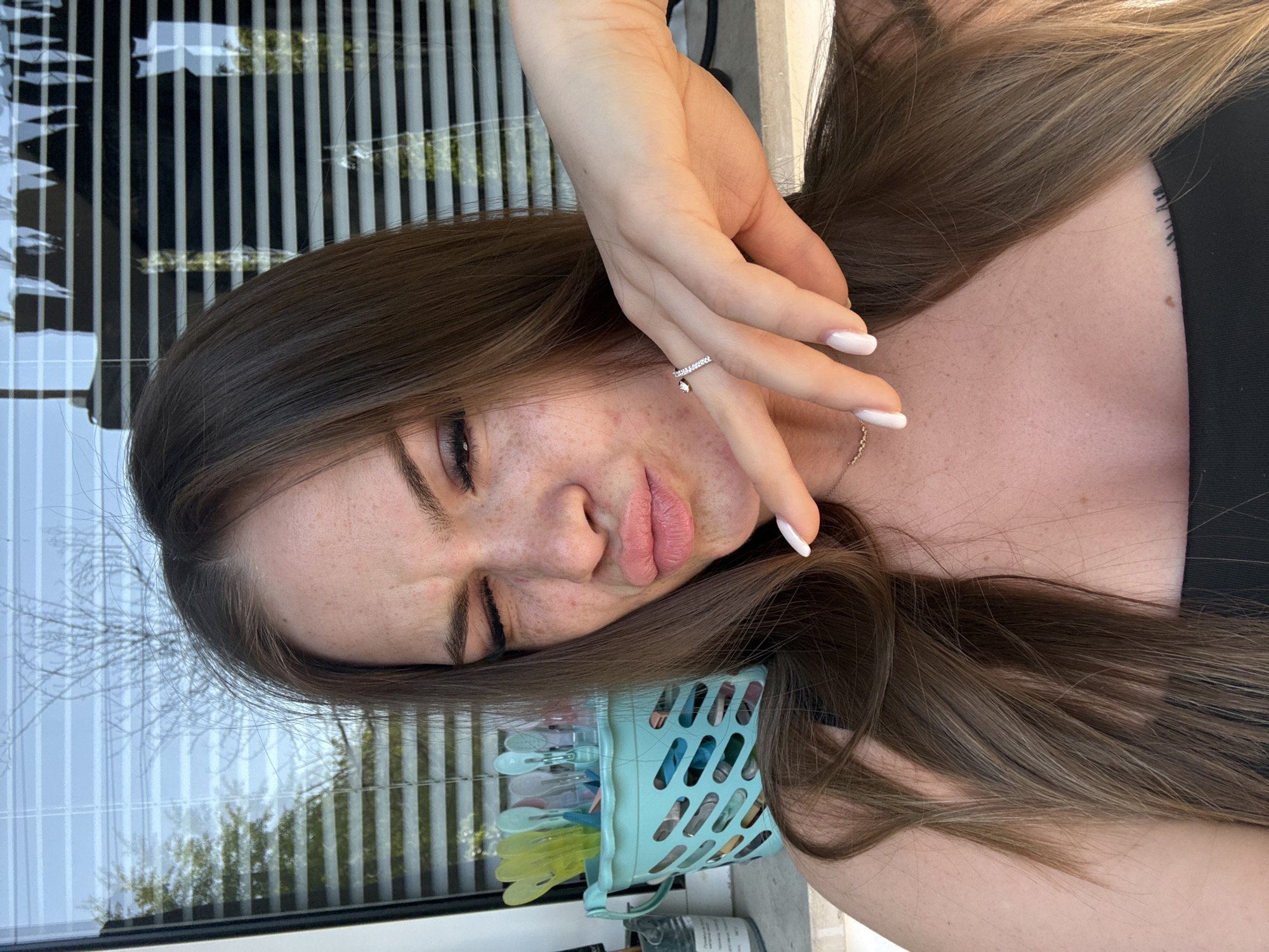 A woman with fair skin and brown hair is posing indoors, holding her hand near her face in a playful gesture. She has freckles, and is wearing a ring on her finger. Her nails are painted. She is wearing a black top. Next to her is a blue plastic basket with clothespins inside. Through a window, vertical blinds are visible. The location of the image cannot be determined, therefore the location in the city of Plovdiv, Bulgaria is unknown.