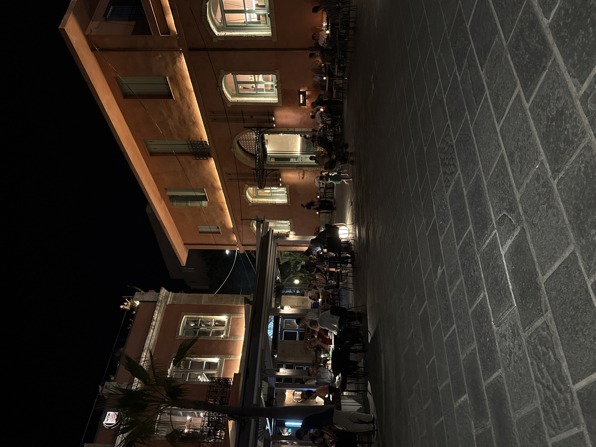 The image depicts an outdoor dining scene at night in an urban environment. Two buildings with orange facades are visible, illuminated by warm lighting. The building on the left features a palm tree in front and a rooftop terrace with several people. The building on the right has large arched windows and a sign that reads "TATAKI". Numerous tables and chairs are set up on a stone-paved plaza, with many patrons seated and dining. The atmosphere appears lively, with some people walking and interacting. The overall scene suggests a restaurant or a row of restaurants in a city square at night.