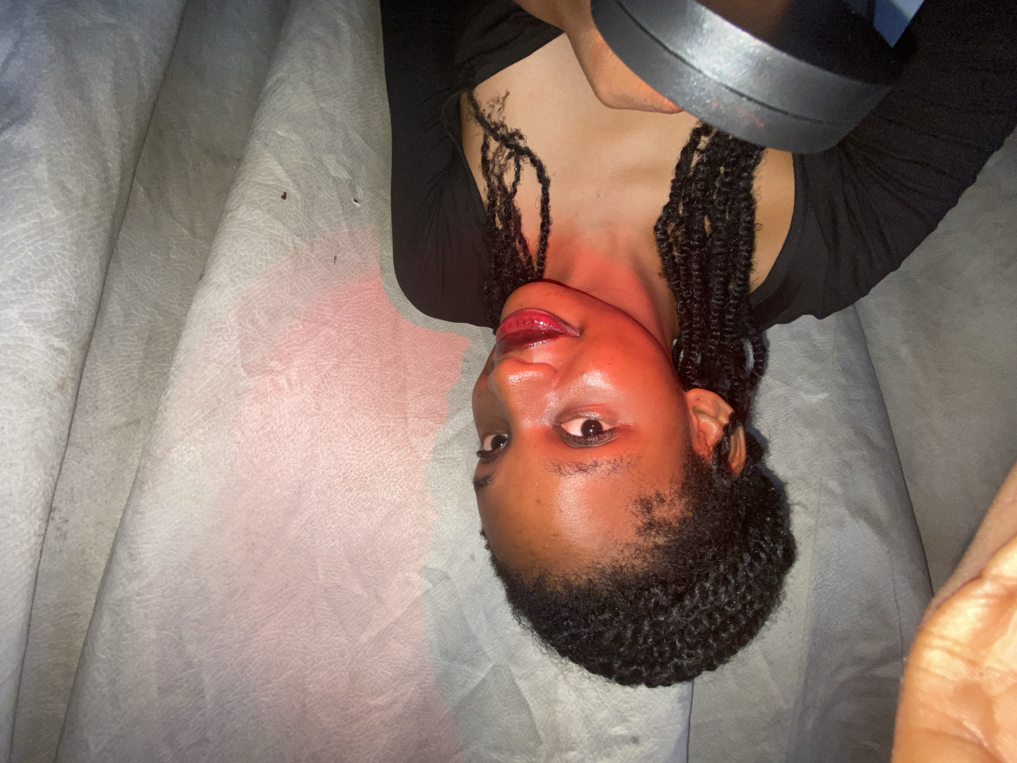 A person lies on a pale gray textured surface, possibly a bed or couch. The person's face is illuminated with a strong red light source, casting dramatic shadows. The person has dark skin, glossy lips, and dark eyes that look towards the left. The person's hair is styled in dark braids. The person is wearing a dark, possibly black, top. A black object, perhaps a light fixture, is positioned above the person. Location: Unknown, Unknown.