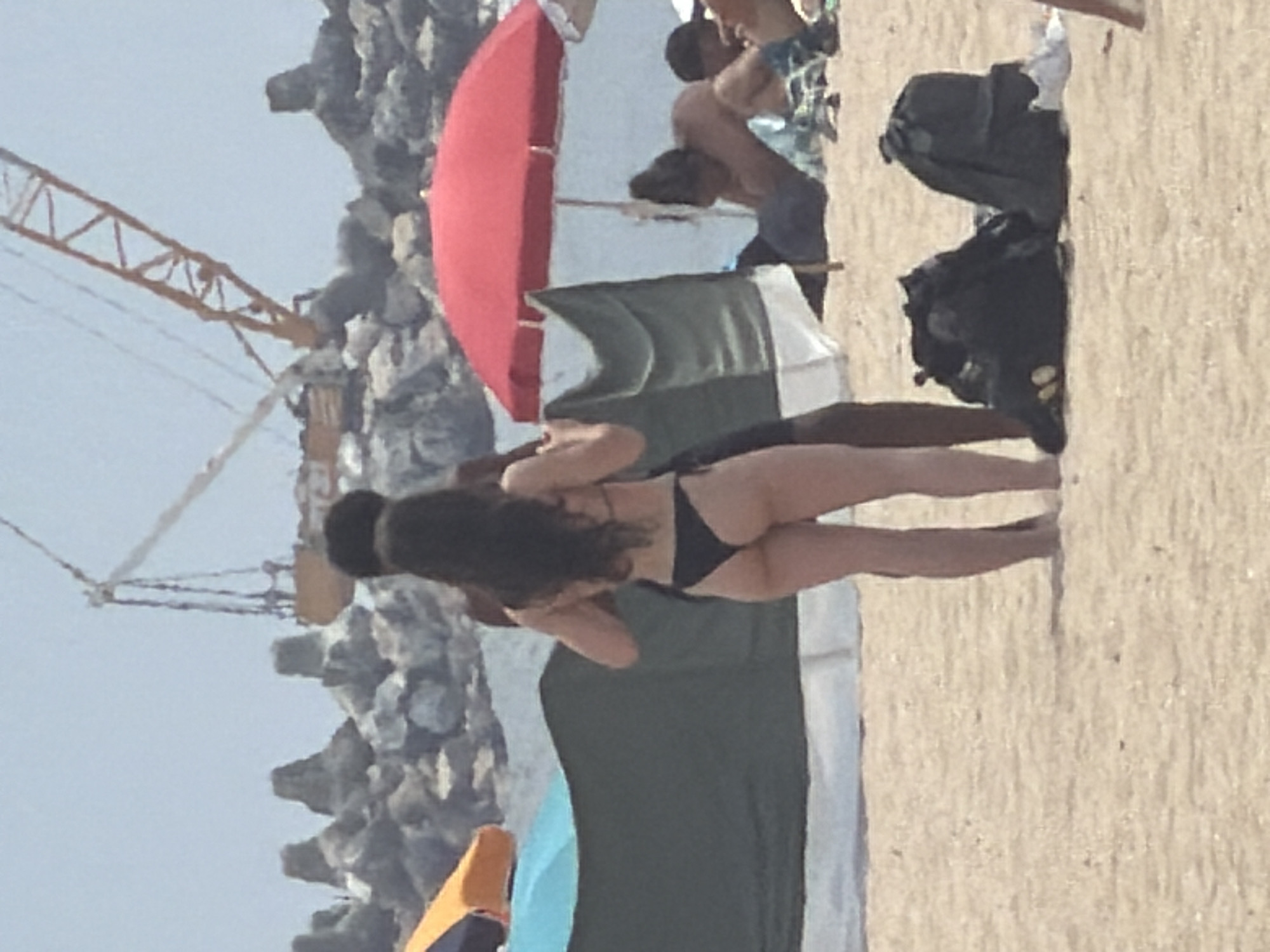 This is a photograph taken at the Praia da Baía in Espinho, Portugal. In the foreground, a woman in a black bikini is seen from the back, standing on the sand. To her left, a man's torso is visible. Behind them, a large green and white windbreak is set up. In the background, a red beach umbrella provides shade for people sitting on the sand. Further in the distance, a construction crane is visible against the sky, along with rock formations and the ocean. Two black backpacks are situated on the sand in the lower right portion of the image.