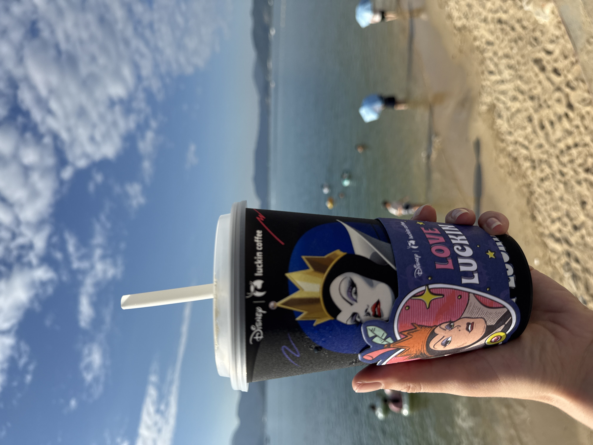 A person's hand is holding a black paper coffee cup with a white lid and straw. The cup is decorated with Disney characters and the Luckin Coffee logo. The background shows a beach with people, umbrellas, and the ocean under a blue sky with scattered clouds. The location is Longgang District, China.