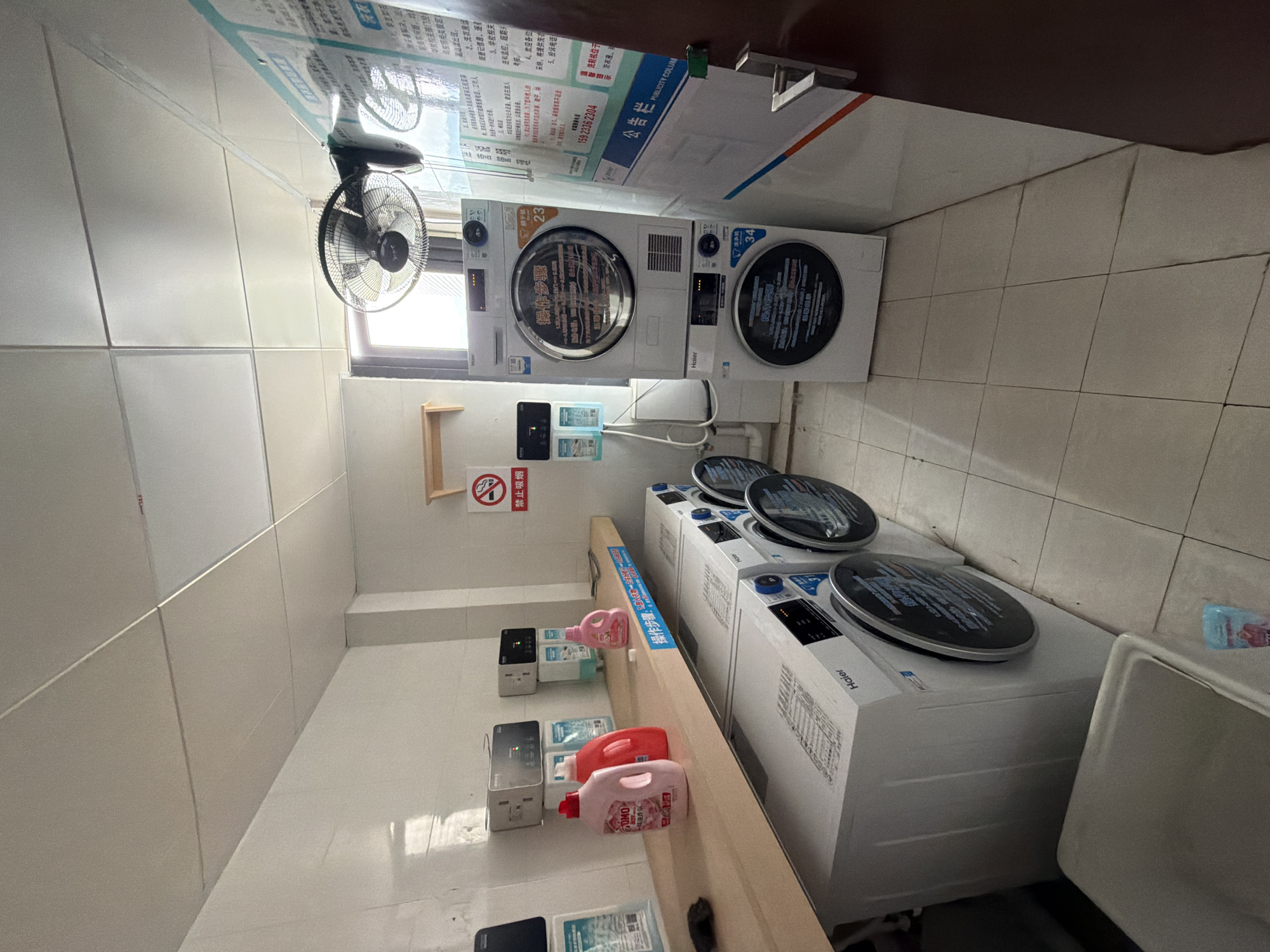 The image depicts a laundry room in Jiangjin, China. The room contains several washing machines and dryers, a shelf with laundry detergent bottles, and informational posters on the wall. The overall lighting is artificial, with fluorescent lights on the ceiling. The room appears to be tidy and functional, suggesting it is a common area for residents or customers. A fan is mounted on the wall near a window, likely for ventilation. The washing machines are branded Haier. One dryer is stacked on top of another washing machine. There is a white laundry sink in the foreground.