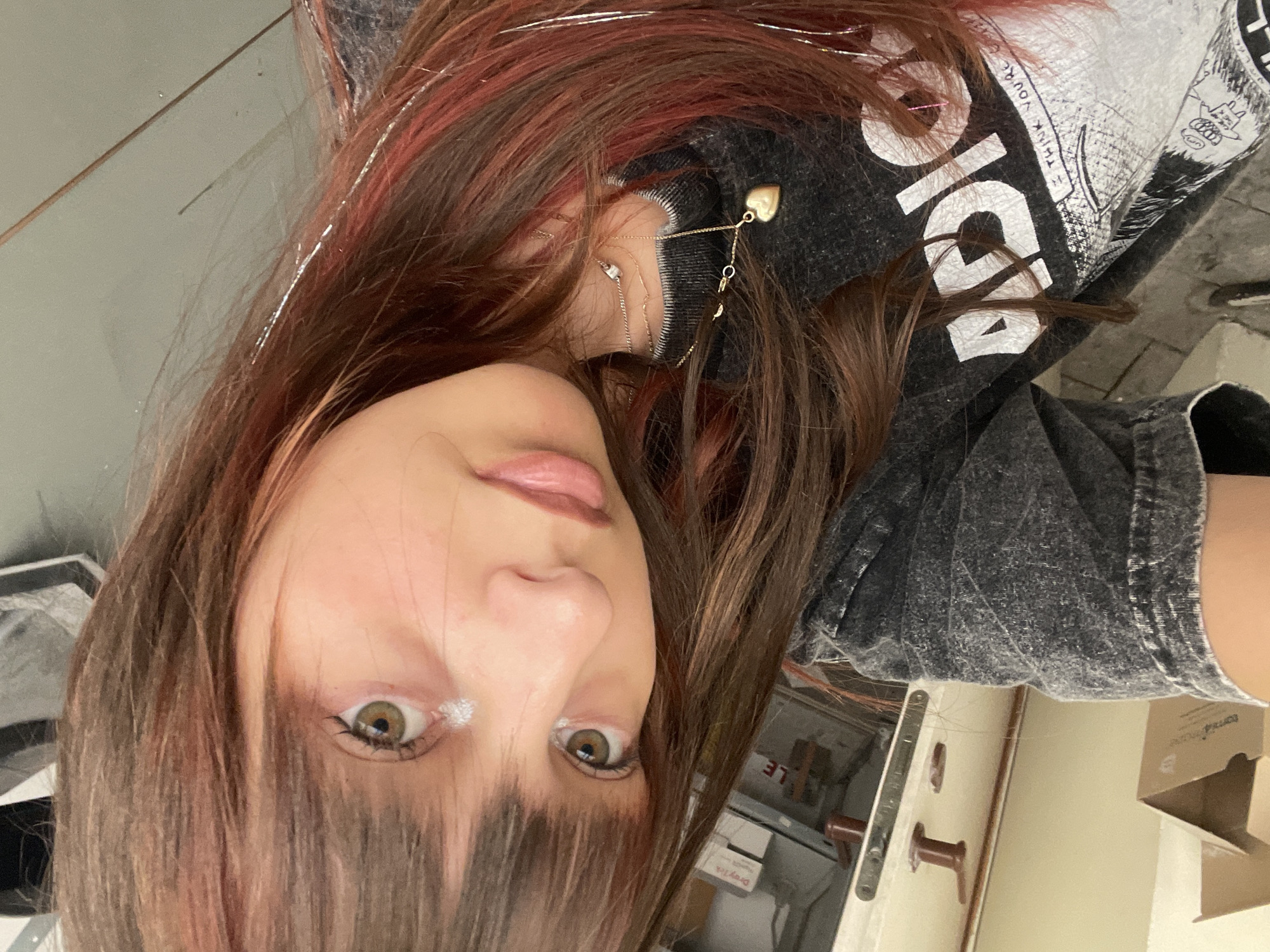 The image shows a young woman with reddish-brown hair and bangs, wearing a grey t-shirt. She has a heart-shaped pendant necklace. Her eyes are open and her head is tilted. There is some sparkly material in her hair. The background is blurred and appears to be an indoor setting, possibly a home or studio. There are some boxes and furniture visible in the background. The overall lighting is dim. No specific location in Raanana, Israel is identifiable from the image.