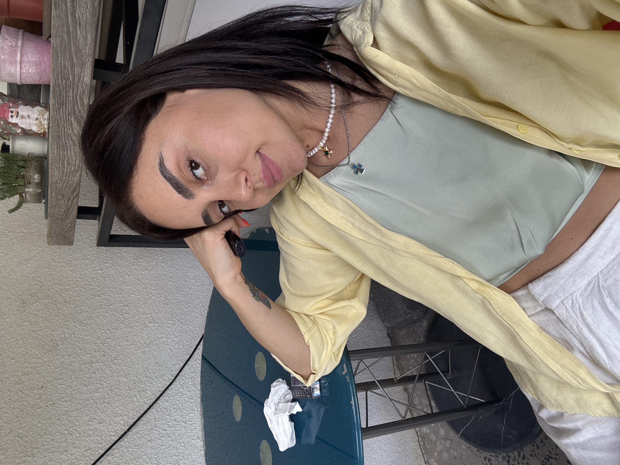 A young woman with dark hair, wearing a light yellow unbuttoned shirt over a light green camisole and white pants, is sitting at a teal colored table on what appears to be a balcony. She is resting her head on her hand, looking towards the camera with a slight smile. On the table, there is a crumpled white napkin and a pack of cigarettes. In the background, there are some potted plants on a shelf, and the wall has a textured finish. The overall setting suggests an outdoor urban environment in Tbilisi, Georgia.