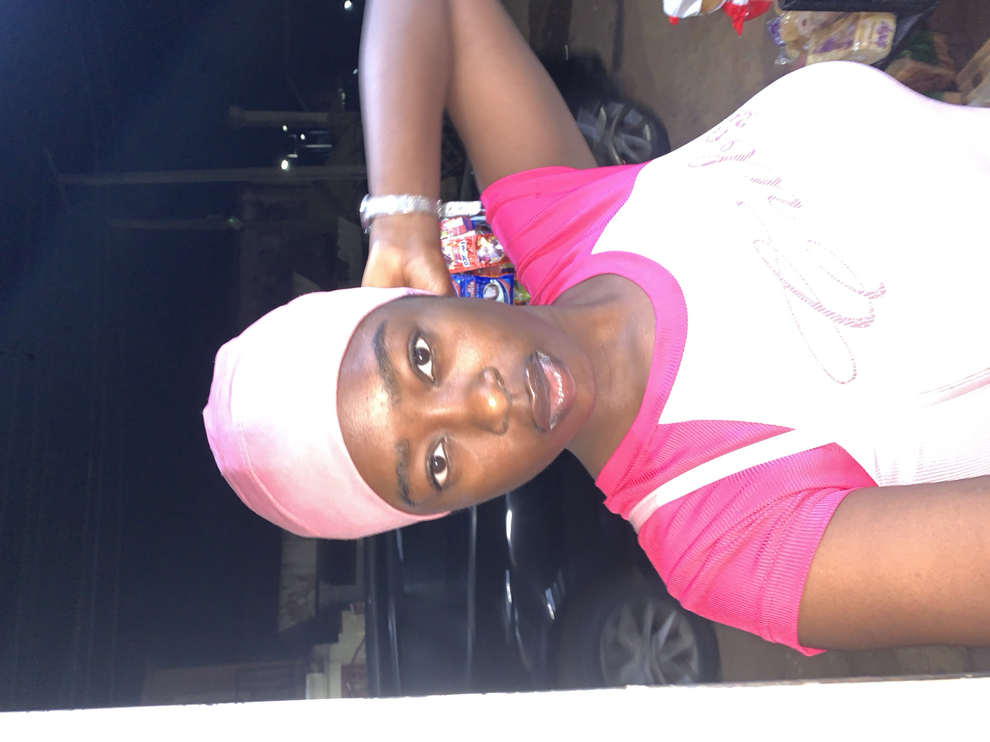 A young, brown-skinned woman wearing a pink headscarf and a white t-shirt with pink sleeves is standing outside at night. She is holding her left hand up to her head. A silver watch is visible on her left wrist. Behind her, there is a black car and a small shop with snacks and other items displayed. The location is the city of New Stadium, Nigeria.