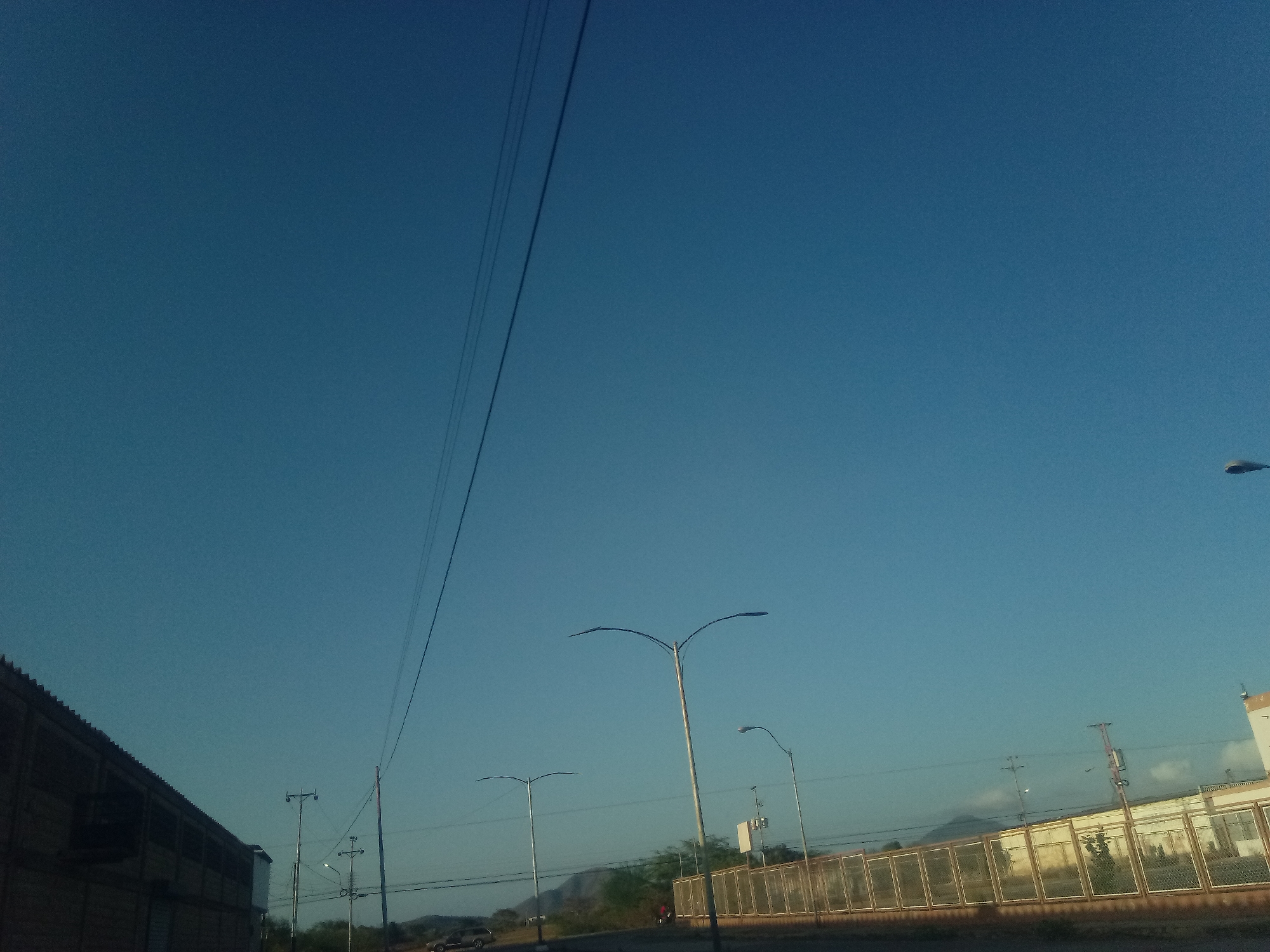 A daytime view of Juan Griego, Venezuela, shows a large building on the left, and a fence on the right. Streetlights line the road. Power lines run above. A car is on the street in the distance. Mountains are on the horizon. The sky is a clear, bright blue.