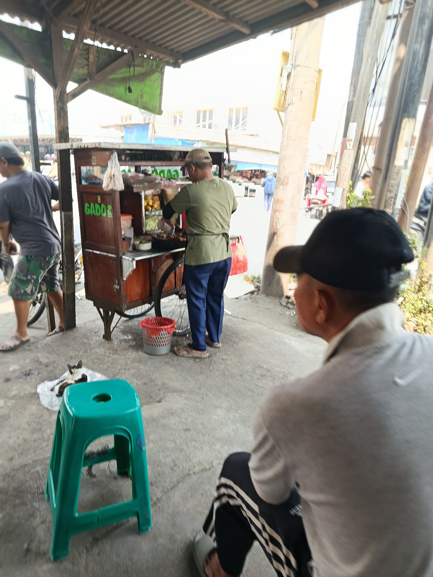 In Wanasari, Indonesia, a street food vendor prepares a dish from a wooden cart labeled "GADO2". The vendor, wearing a green shirt and a cap, is focused on his work. A customer, partially visible from behind and wearing a black cap, sits on the right, facing away from the camera. To the left, another individual in a grey shirt and patterned shorts is also partially visible. In the foreground, a bright green plastic stool sits on the concrete ground, with a cat resting on some white plastic nearby. The scene takes place outdoors under a makeshift shelter with a corrugated metal roof, with utility poles and a blurred background of buildings and traffic visible. The lighting suggests it is daytime, likely morning or afternoon, with bright sunlight creating shadows and highlights.