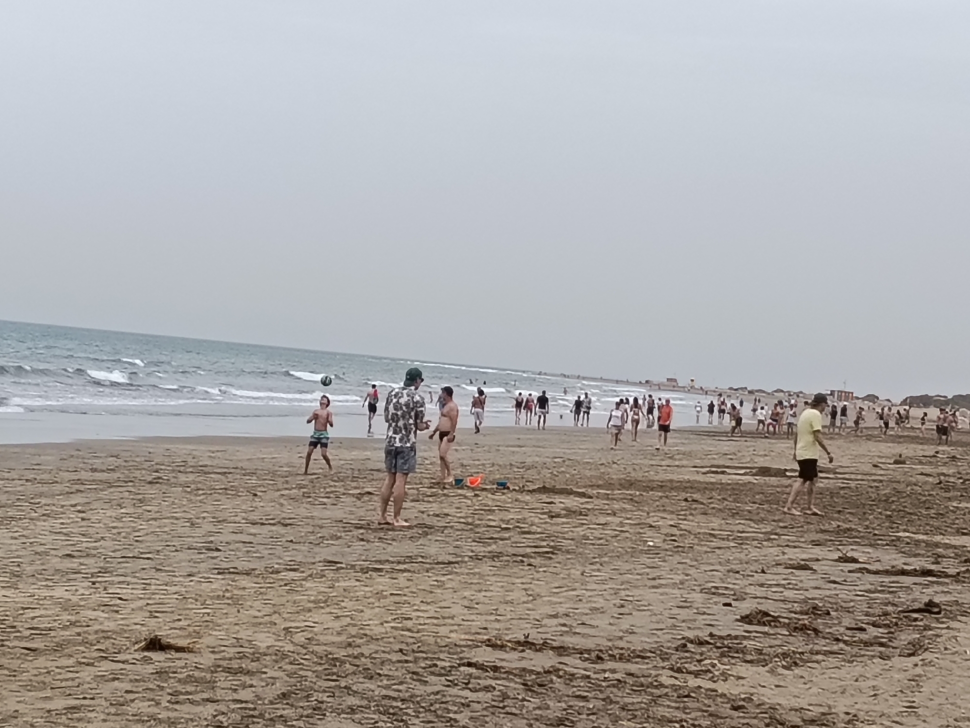 The image captures a wide shot of a crowded beach in San Bartolomé de Tirajana, Spain, under an overcast sky. The scene is dominated by a vast expanse of wet, light brown sand that stretches from the foreground to the distant shoreline.
In the mid-ground, several individuals are engaged in activities. To the left, a young man in dark swim trunks and a boy in patterned swim shorts appear to be playing with a volleyball, which is visible in the air between them. Another man, shirtless and in black swim trunks, stands nearby. Two small, brightly colored plastic buckets, one red and one blue, are left on the sand close to these individuals. Further to the right, a man in a yellow t-shirt and dark shorts walks across the sand.
Along the water's edge, numerous people are scattered, walking, standing, and enjoying the beach. They are seen as smaller figures due to the distance, dressed in various beachwear. The ocean stretches across the left half of the background, with gentle white waves breaking along the shore. The sky is a uniform, pale gray, indicating an overcast day without direct sunlight. On the far right horizon, faint structures or buildings suggest a developed coastal area. No visible text is discernible in the image.