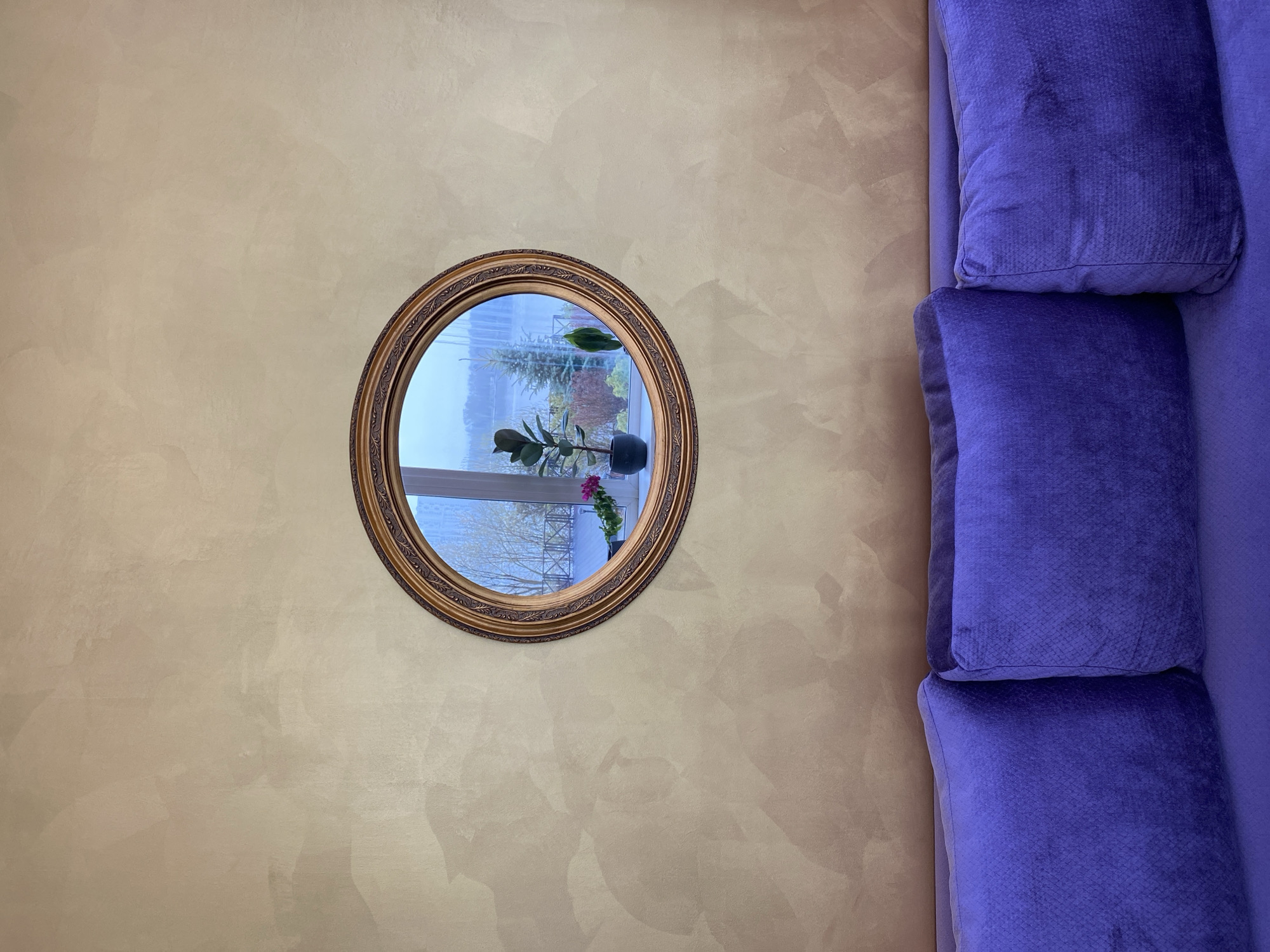 A close-up shot depicts an interior scene with a combination of beige wall texture and a section of a purple sofa. A round mirror, framed with ornate gold trim, is mounted on the beige wall, reflecting an outdoor scene with a plant on a windowsill, a view of buildings and trees under a pale sky. The purple sofa segment on the right comprises three cushions, each displaying a slightly textured fabric. The overall scene is well-lit. While the reflection in the mirror alludes to an external environment, no specific details confirm the location to be Yekaterinburg, Russia.
