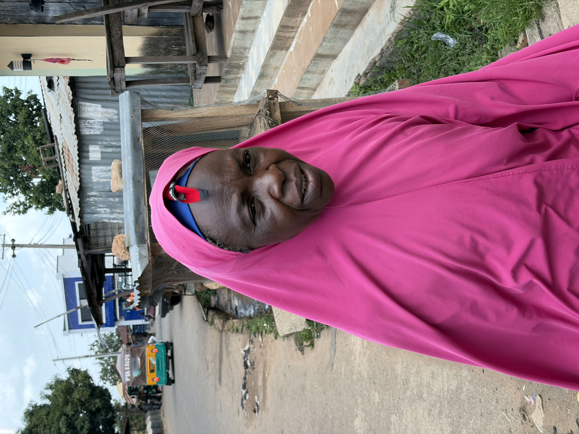 A person wearing a bright pink hijab stands outside on a street. A blue underscarf is visible. A red and silver decorative pin is affixed to the side of the hijab near the face. In the background, there is a dusty street, buildings, trees, and a vehicle. The location appears to be in the city of Jos, Nigeria.