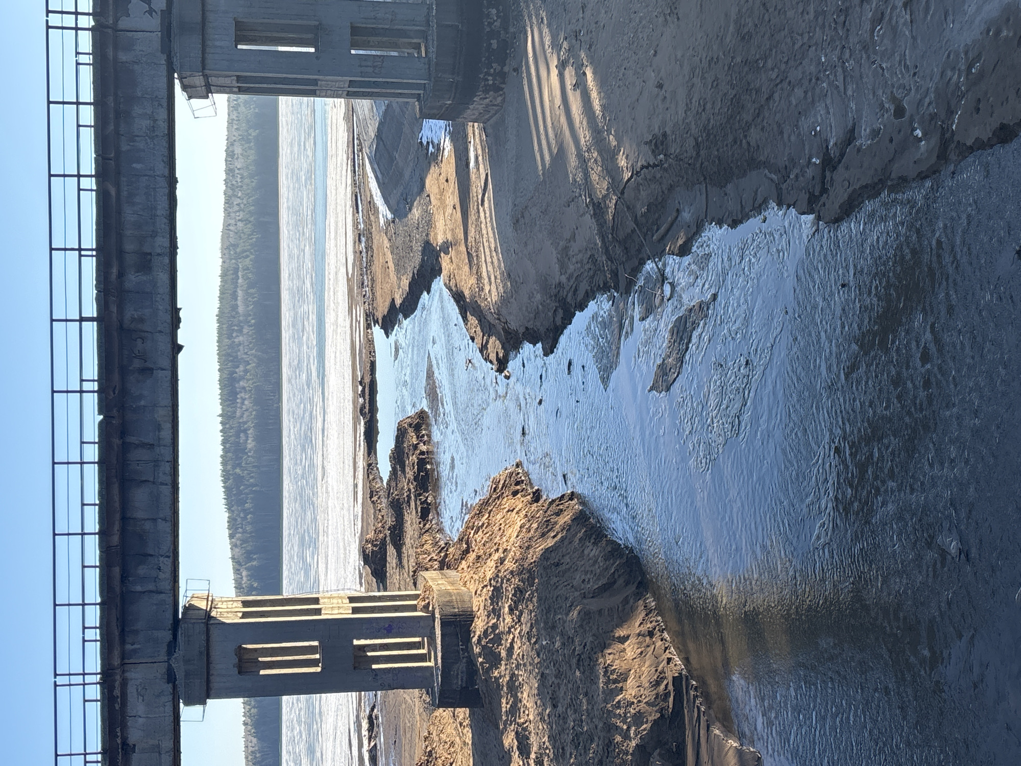This photograph captures an industrial scene in Chaikovsky, Russia. The prominent features include a dilapidated concrete structure with a metal railing, likely part of an abandoned or decaying bridge. Eroded soil and sandbanks line the foreground, with a stream of water flowing between them. In the background, a large body of water reflects sunlight, with a dense forest visible on the distant shore. The sky is clear and blue. The overall atmosphere conveys decay and environmental degradation.
