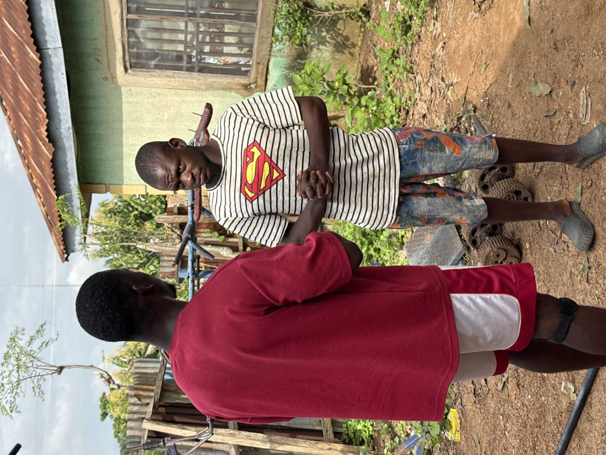 In Akwanga, Nigeria, two young Black men are engaged in conversation outdoors. The man on the left, facing away from the camera, wears a red t-shirt and white and red shorts. The man on the right, facing the camera, sports a striped t-shirt with a Superman logo and patterned shorts. They stand in a dusty, outdoor area with a weathered building in the background, featuring a corrugated metal roof and a barred window. Green foliage and a rickety wooden fence are also visible, suggesting a somewhat rural or informal setting within the city. The ground is covered in dirt and scattered debris, including what appear to be old mechanical parts. The sky is partly cloudy.