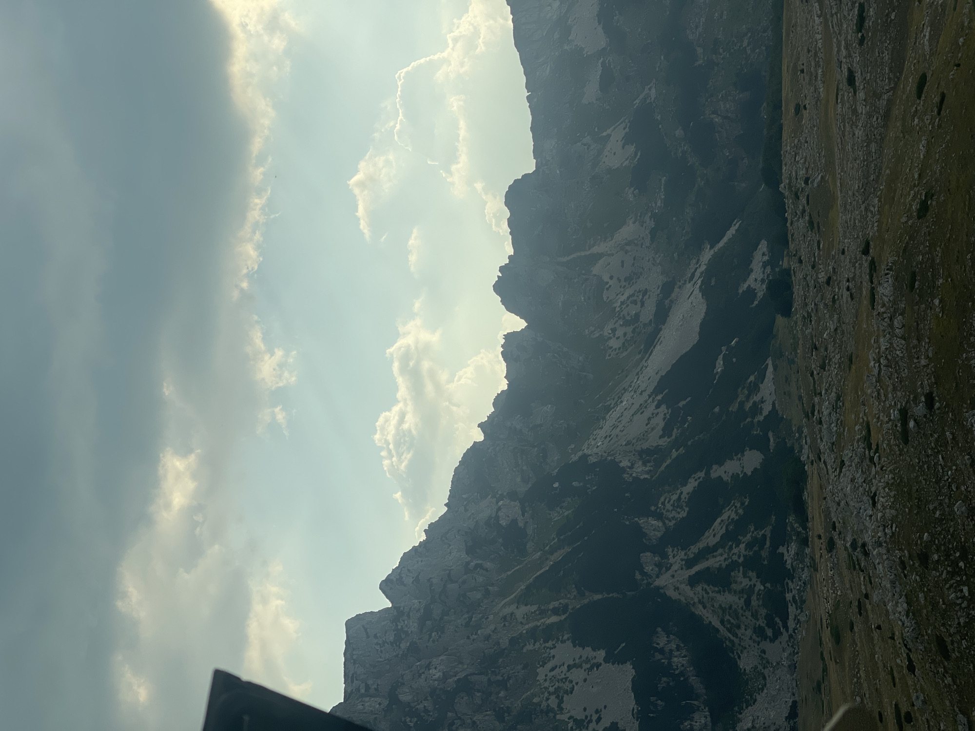 The image depicts a panoramic view of a mountainous landscape. The foreground is a rocky, sparsely vegetated terrain with scattered stones and patches of dry grass. A darker, more densely vegetated area can be seen in the mid-ground, leading into the base of a large mountain range. The mountains themselves are rugged and steep, with visible rock formations and shadows indicating their imposing size. The slopes are a mix of dark green vegetation and lighter rocky scree. The sky above is dramatic, with a gradient of blues and grays, punctuated by bright, illuminated clouds. Sun rays pierce through some of the clouds, creating beams of light that illuminate parts of the sky and the upper mountain peaks. The scene is set in Pašina Voda, Montenegro.