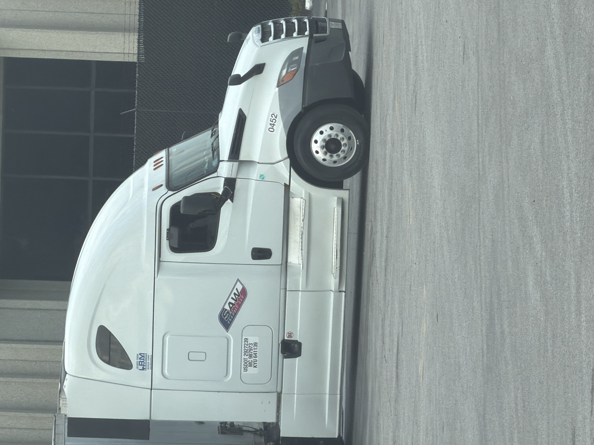 A white Freightliner semi-truck is parked in a lot. The truck has a "SAW TRANSPORT LLC" logo on its side, along with its USDOT number, MC number, and KYU number. The truck is also leased from "LBM LEASING." The scene appears to be in an industrial area, possibly a truck yard, and the background includes a building with large windows and a fence. There are no people visible in the image. The image does not contain any information to indicate its location in Chattanooga, United States of America.